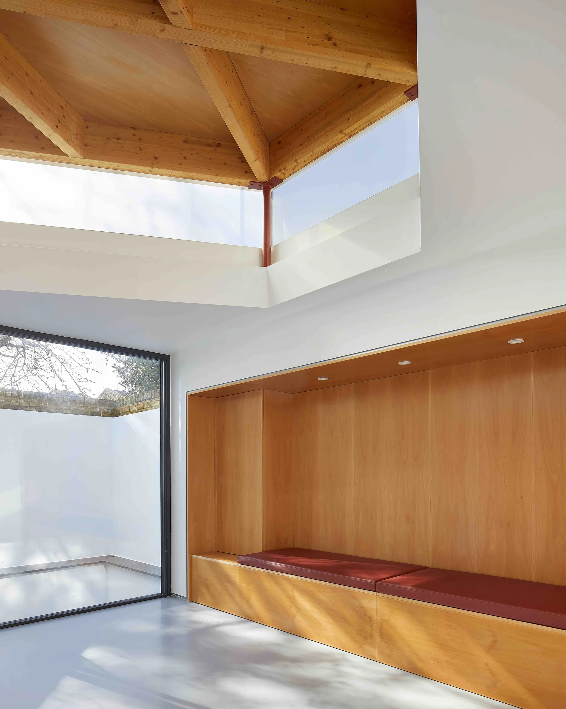 A minimalist interior featuring a built-in wooden alcove bench with red cushions beneath an exposed timber ceiling with clerestory windows.