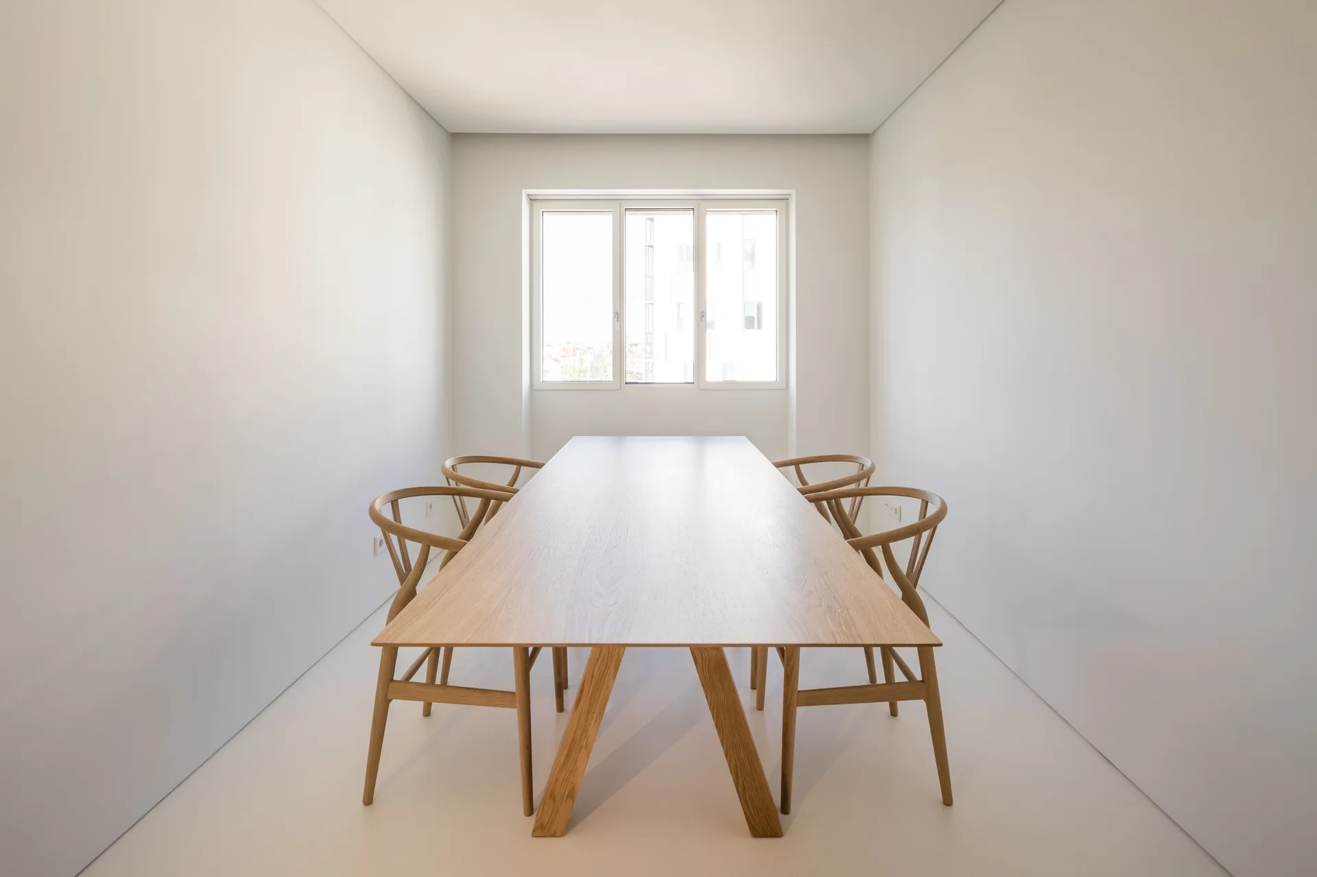 A light wood dining table with four matching wishbone chairs stands in a minimalist room with white walls, a white floor, and a centered window.