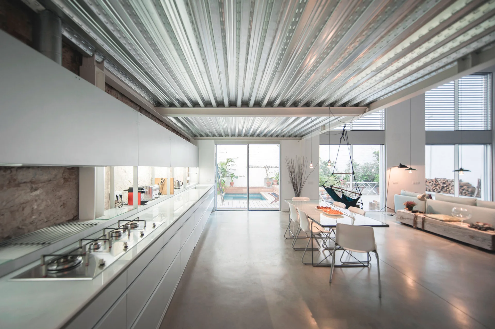 An industrial kitchen and dining space featuring a corrugated metal ceiling, white minimalist cabinetry, a glass dining table, and concrete floors.