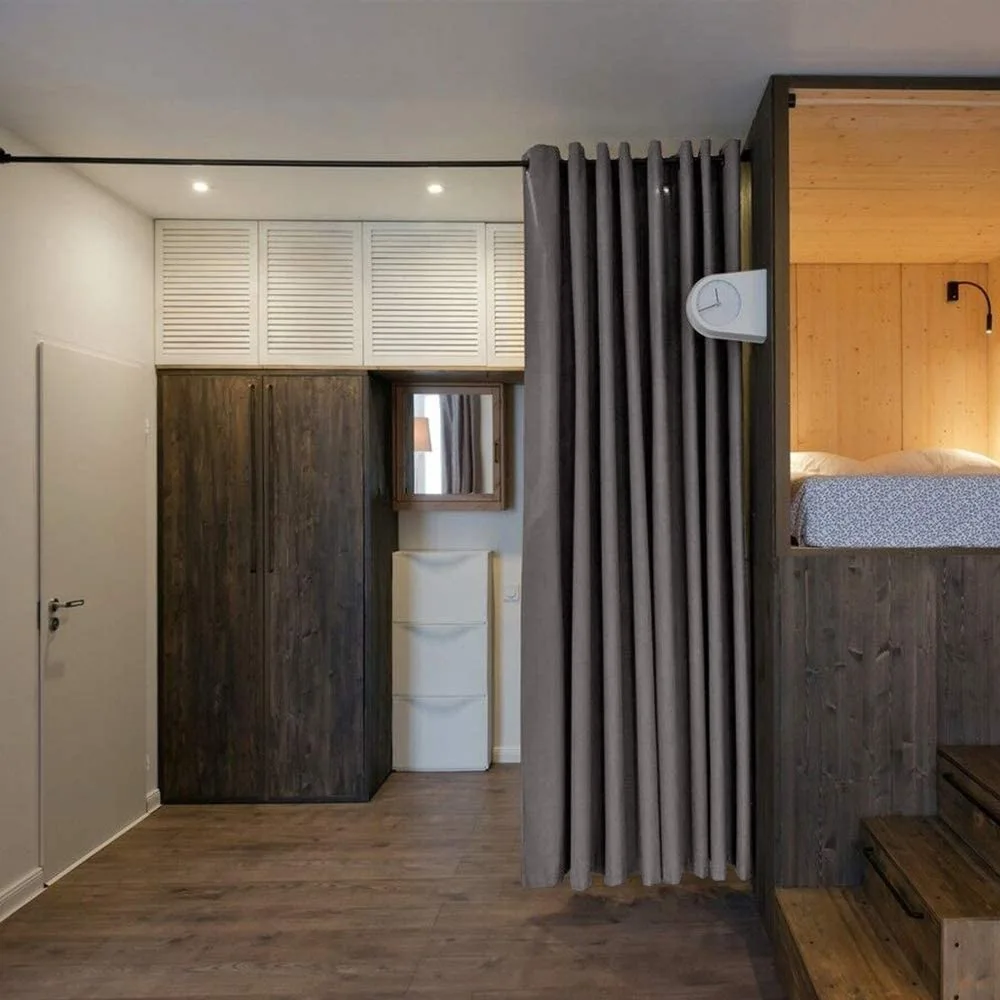 A small bedroom area featuring an elevated wooden loft bed, a grey partition curtain, dark wood wardrobe cabinets, and louvered upper storage.