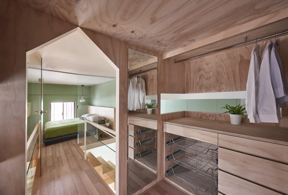 A walk-in closet lined in light plywood features built-in drawers, a mirror, and a clear glass partition looking into a green bedroom.