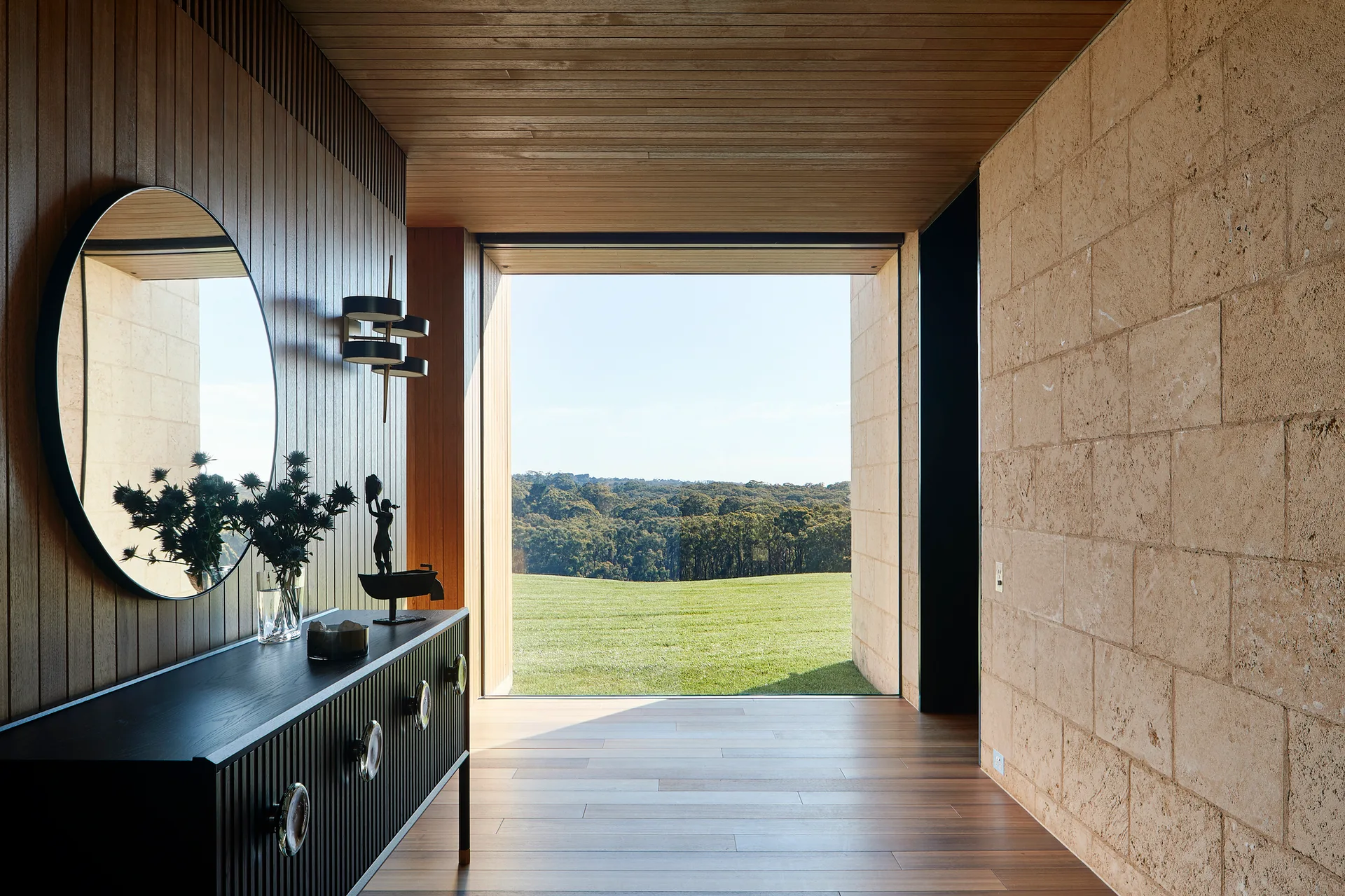 A modern entryway corridor featuring a black console table, a round mirror, a textured stone wall, and a large picture window showing a grassy field.