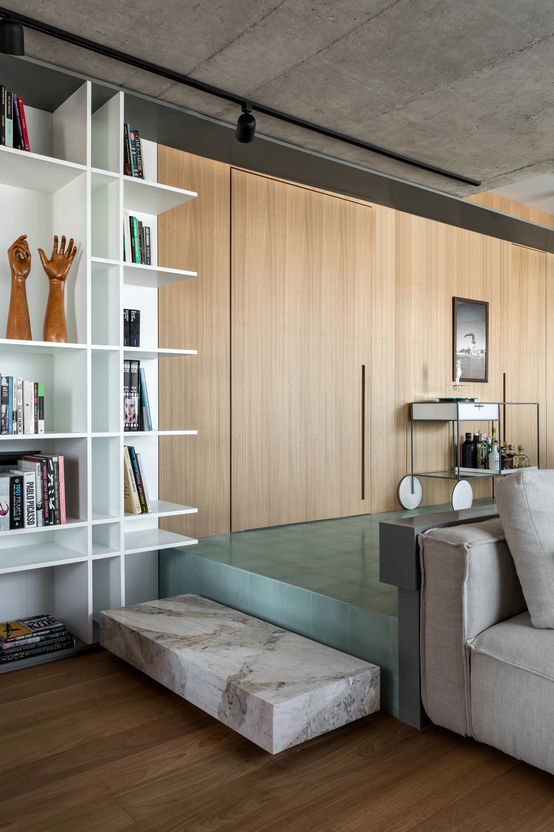 A modern living space featuring a white grid bookshelf, a floating marble step leading to a green tiled platform, and light wood wall paneling.