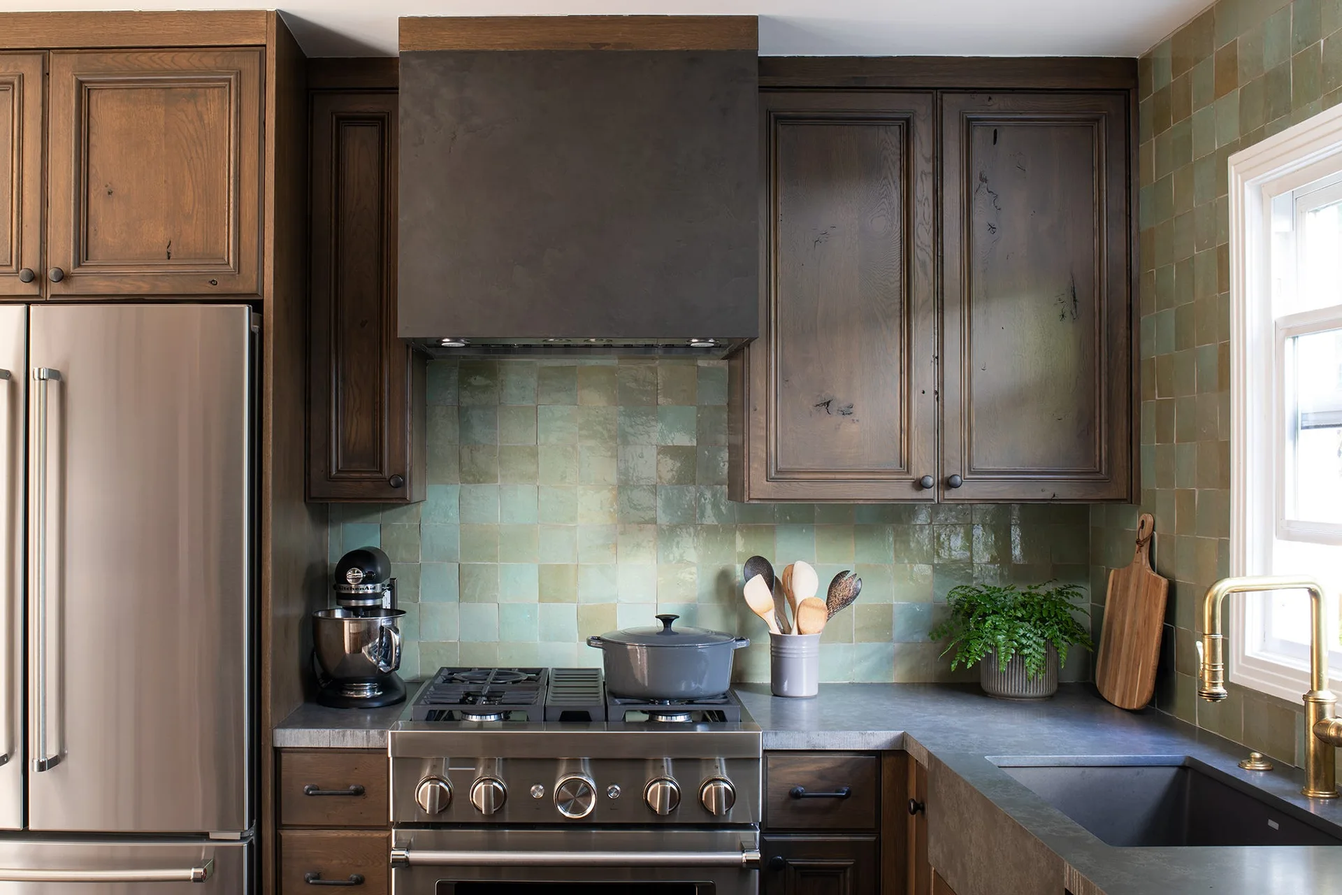 A kitchen features dark stained wood cabinets, a glossy green tile backsplash, a stainless steel range, and a dark matte range hood.