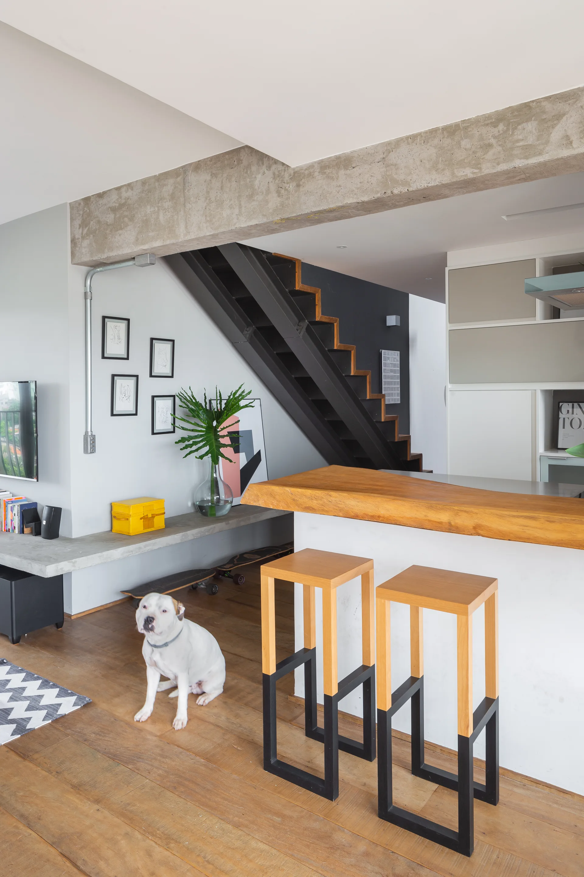 An industrial kitchen and living space features a raw concrete beam, wood-topped breakfast bar with two wooden stools, and a black metal staircase.