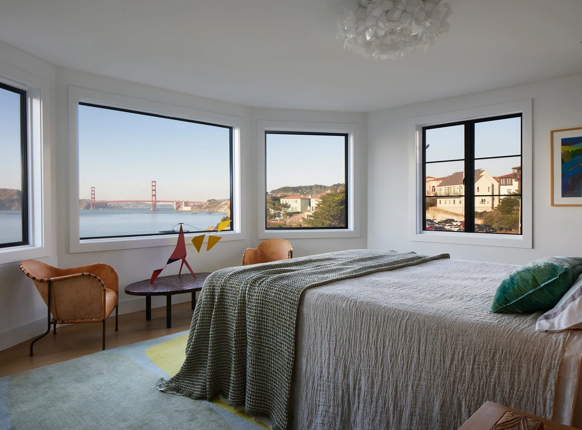 A bright bedroom featuring a bed with textured linens, two tan leather chairs, and picture windows overlooking the Golden Gate Bridge.