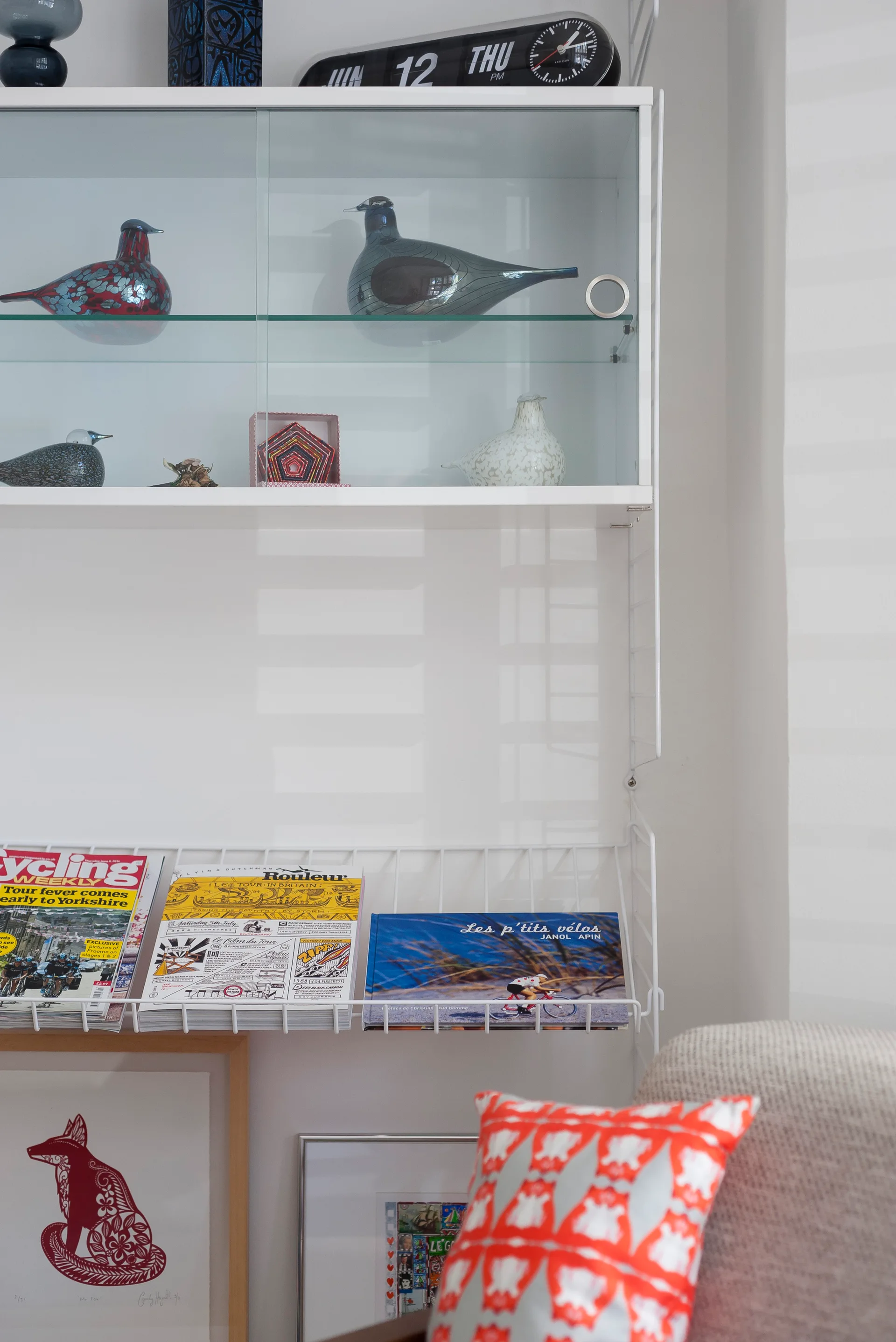 White modular wire shelving on a wall displays glass bird sculptures in a cabinet, cycling magazines on a rack, and a patterned orange cushion below.