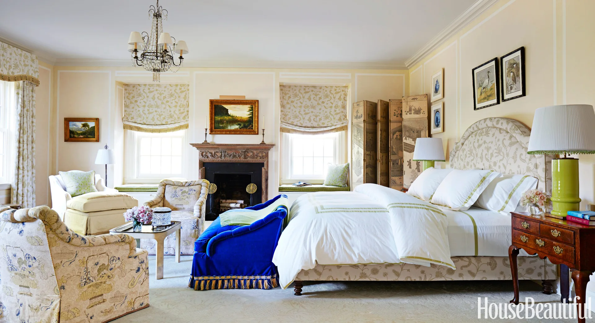 A traditional bedroom features a patterned upholstered bed, a decorative fireplace, and a vibrant cobalt blue velvet bench.