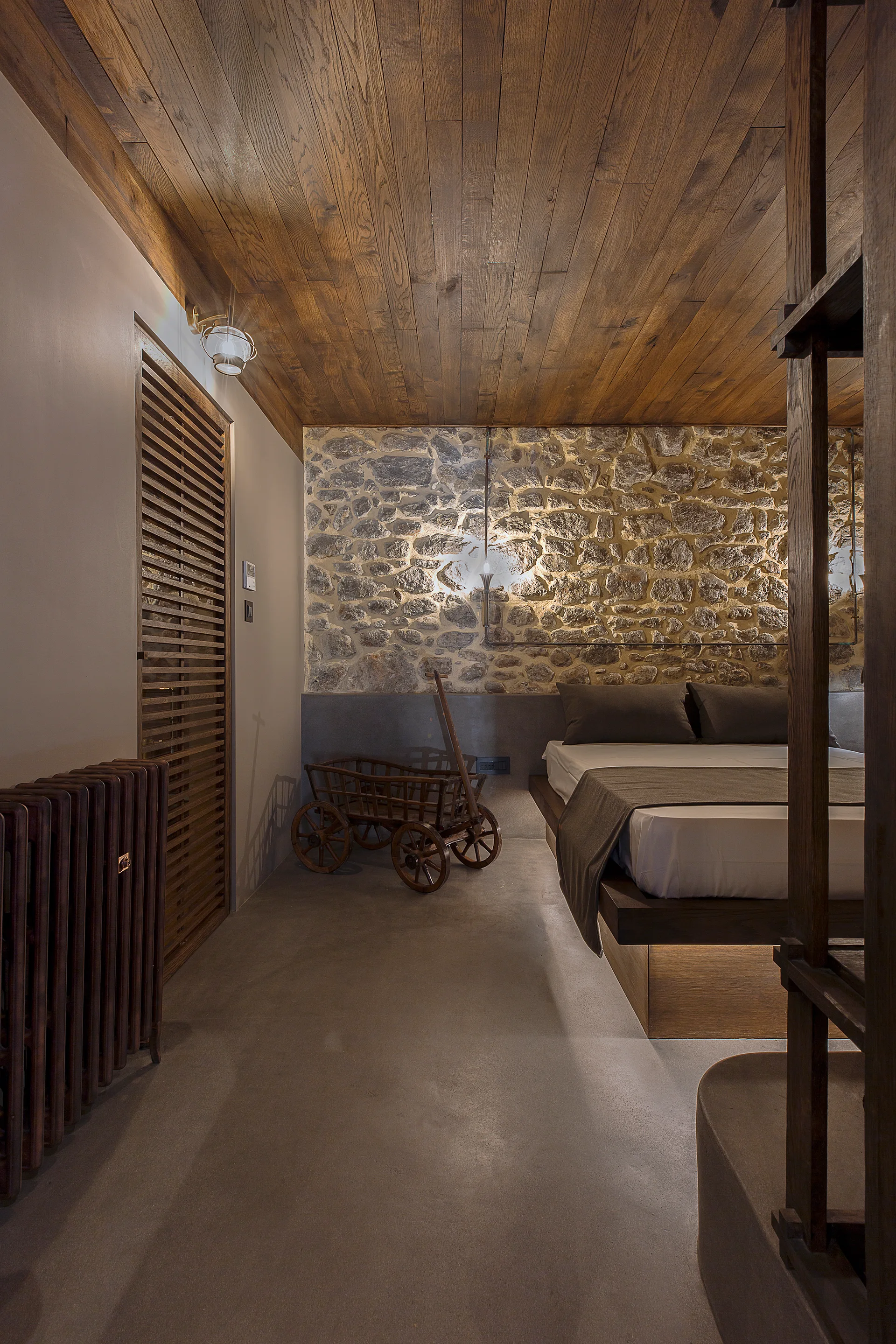 A rustic bedroom features a platform bed against a textured stone wall, complemented by a wooden plank ceiling and smooth concrete flooring.