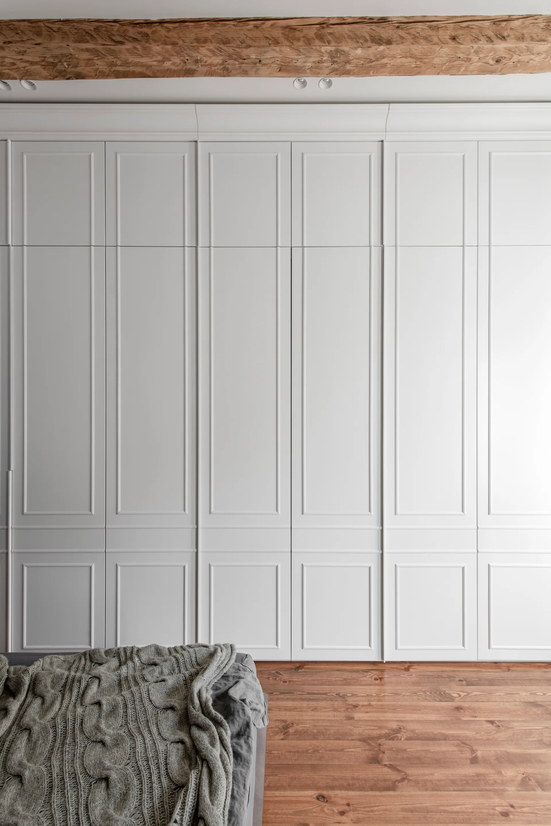 A floor-to-ceiling wall of white paneled built-in wardrobe doors stands above warm wood floors next to a bed with a gray cable knit blanket.
