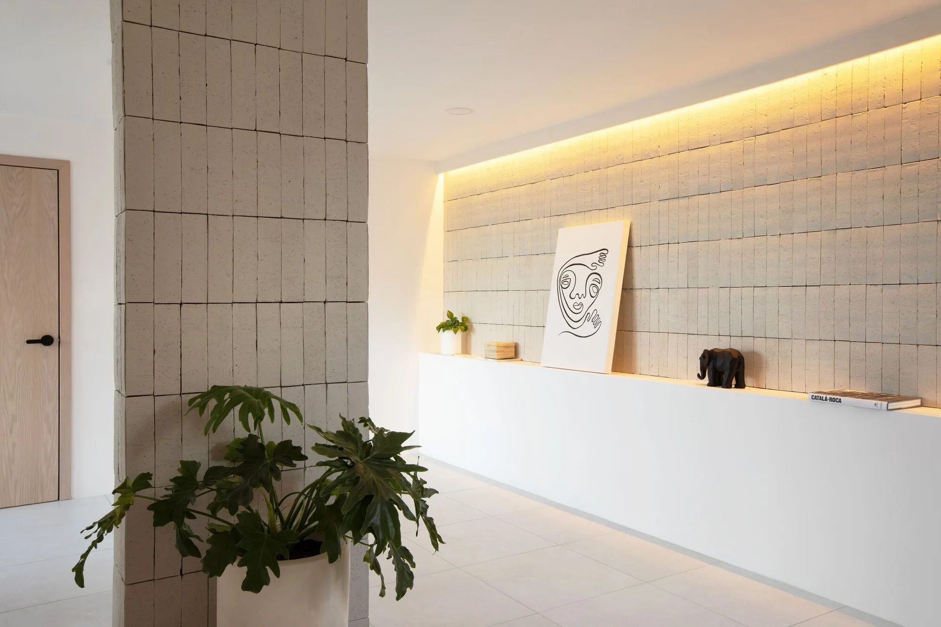 A minimalist hallway features a tiled accent wall with integrated cove lighting, a built-in white console, and a large tile-clad column in the foreground.