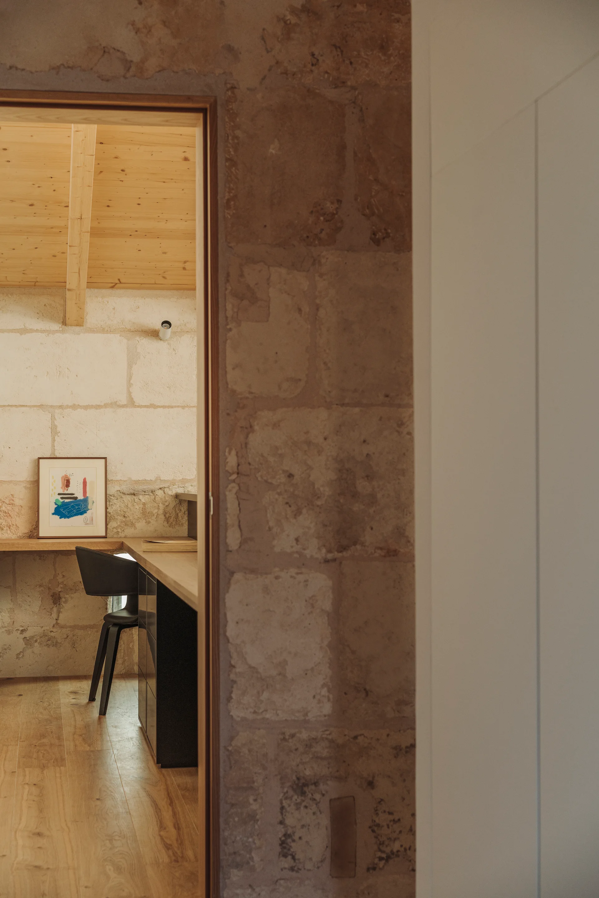 A view through a doorway into a minimalist home office featuring rough-cut stone walls, a built-in wooden desk, and a modern black chair.
