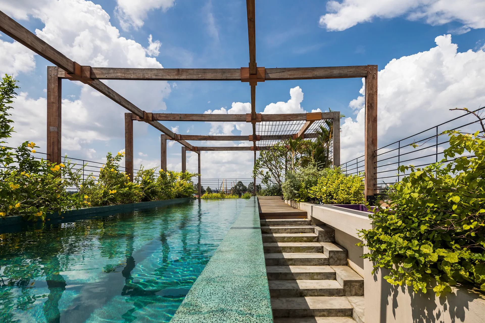 A turquoise rooftop lap pool sits under a large timber pergola alongside concrete steps and lush green landscaping.