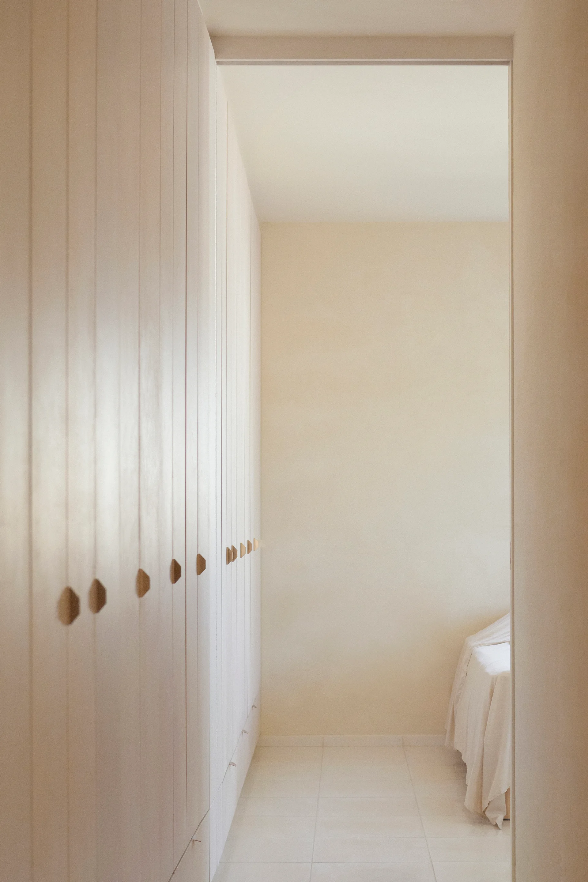 A minimalist dressing corridor features a wall of floor-to-ceiling cream cabinets with small wooden pulls leading to a bedroom.