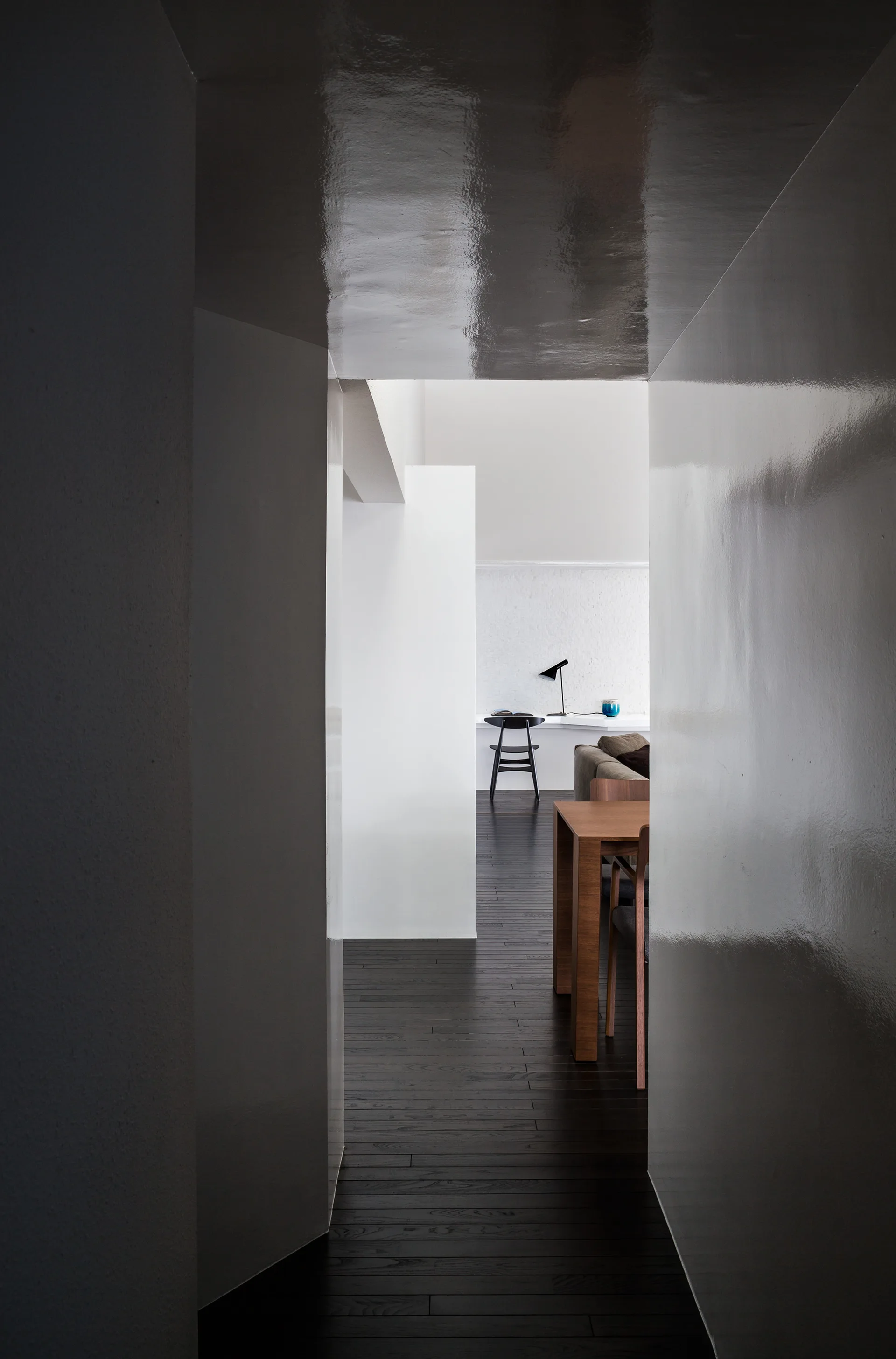 A narrow grey hallway with highly reflective glossy walls looks toward a brighter dining space with dark wood floors.