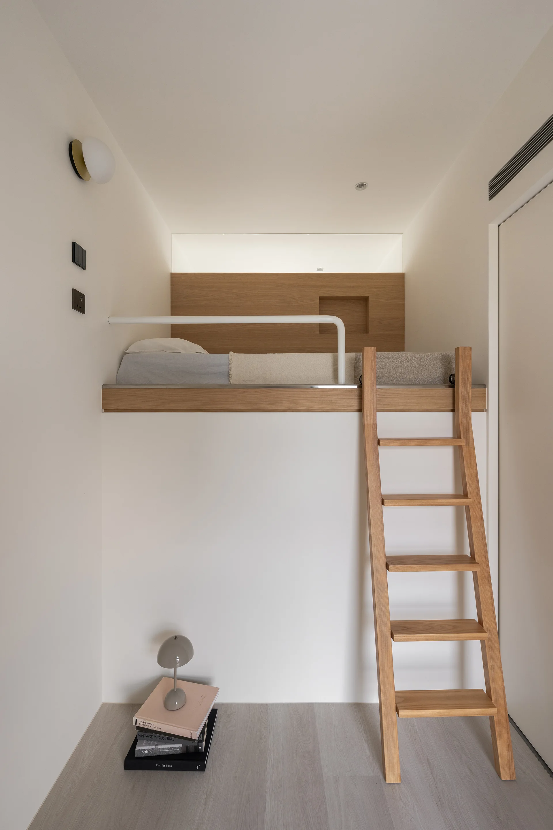 A minimalist white bedroom featuring an elevated built-in loft bed with a wooden ladder, safety rail, and books stacked on the floor.