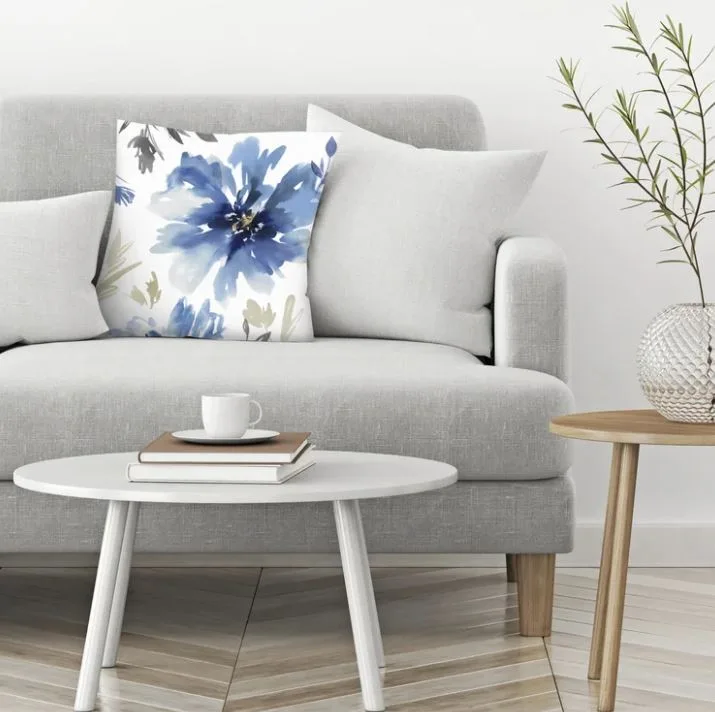 A light gray fabric sofa is decorated with a blue watercolor floral pillow next to a round white coffee table and light wood flooring.