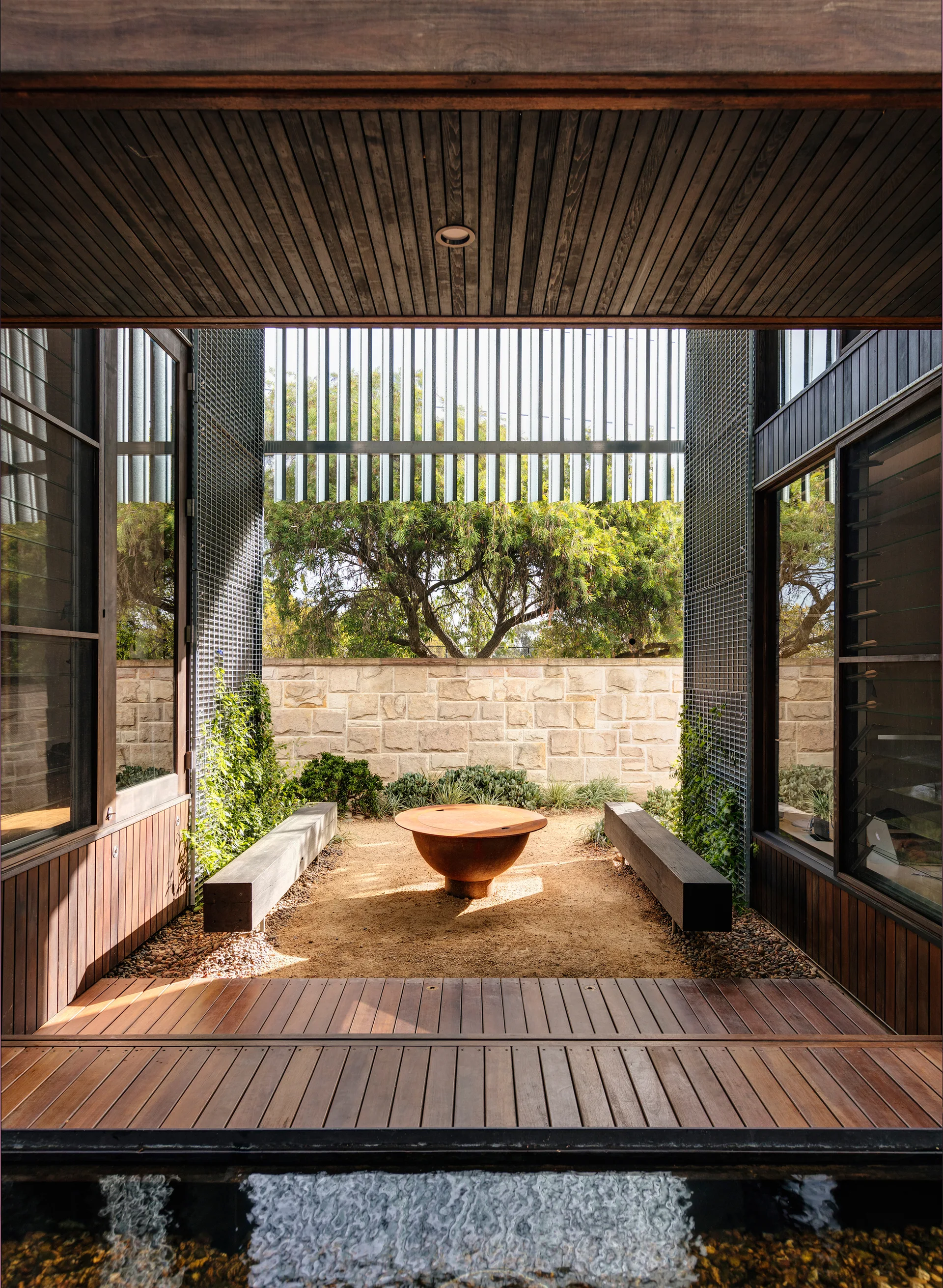 A minimalist outdoor courtyard features a central rusted metal fire pit, two timber benches, a sandy ground, and a light stone wall in the background.