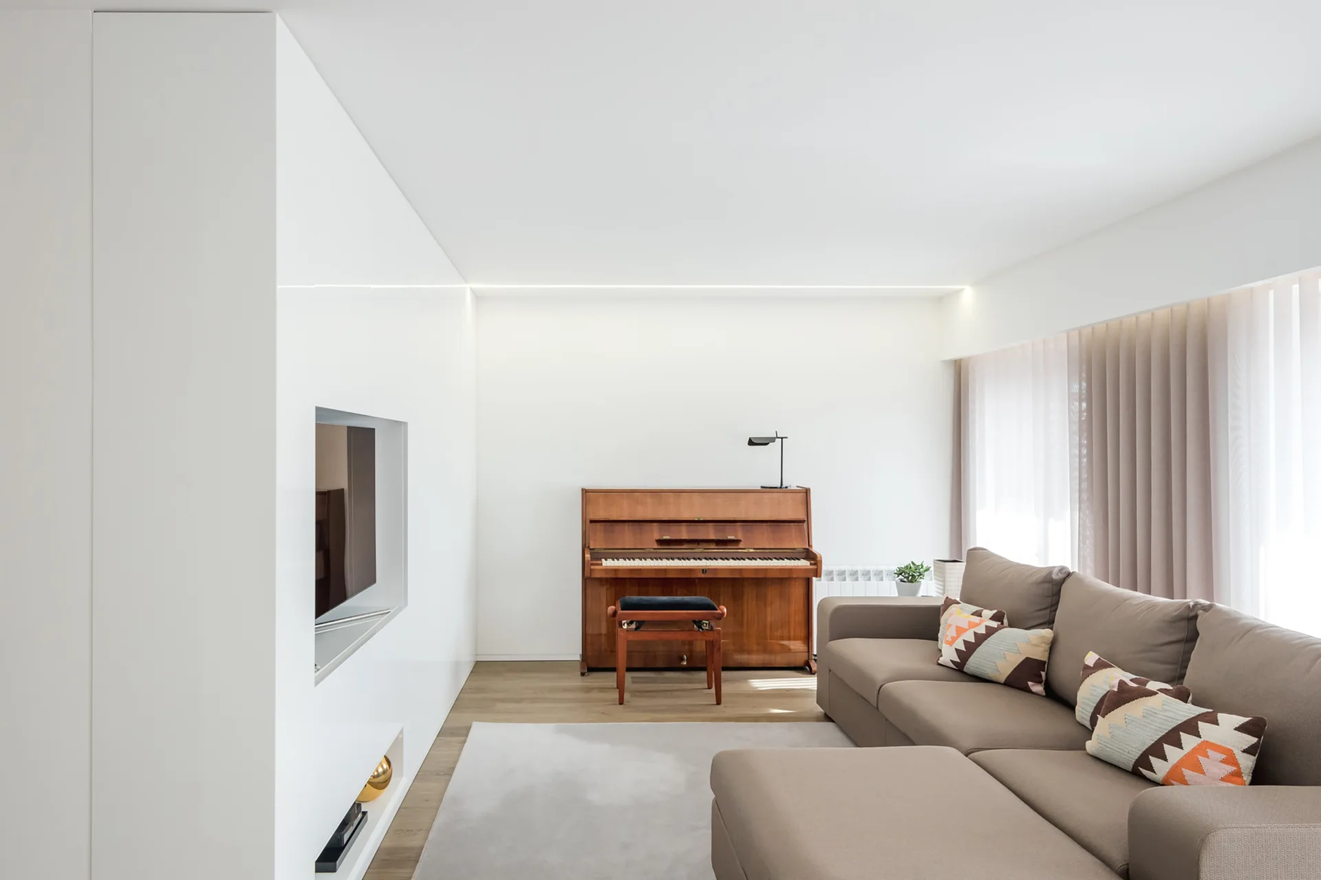 A minimalist living room featuring a tan sectional sofa, a wooden upright piano, and a sleek white built-in media wall with an integrated television.