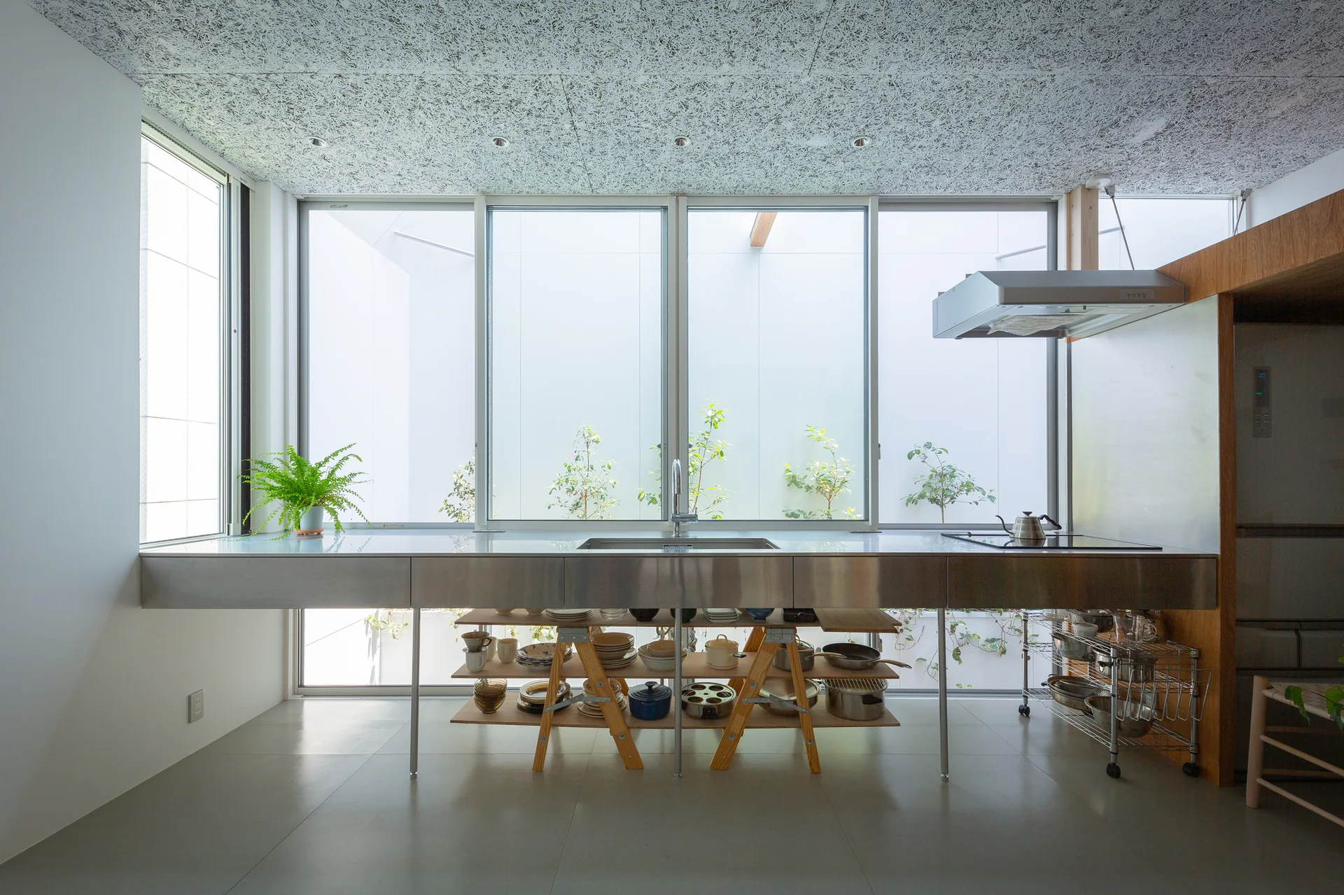 A minimalist kitchen features a long stainless steel counter with an undermount sink, open wooden shelving, and large windows looking out to a courtyard.