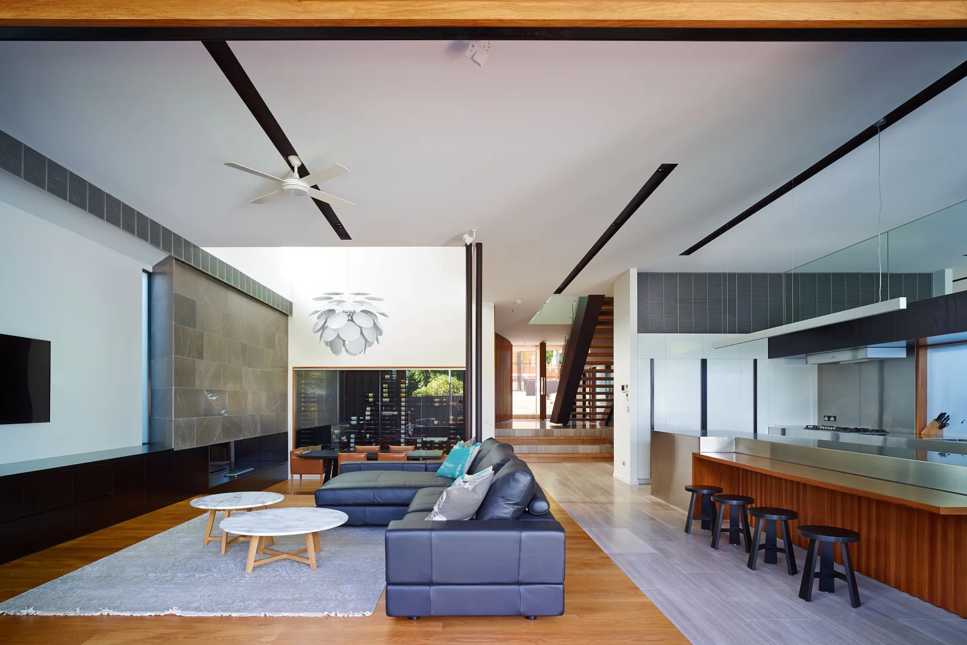 A modern open-plan living room with a black leather sectional sofa, marble-topped coffee tables, a stone fireplace, and a wood kitchen island.