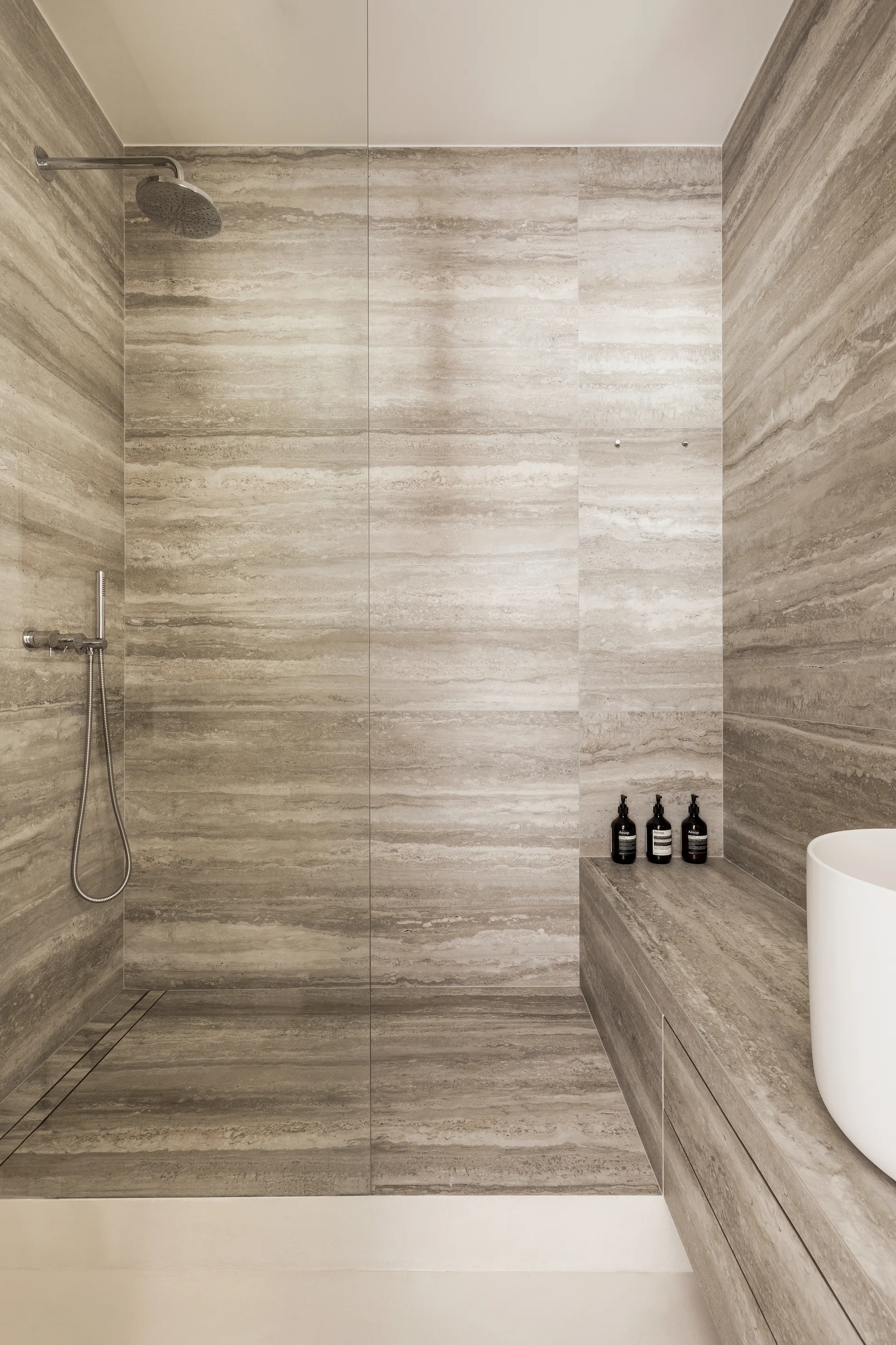 A minimalist walk-in shower features warm grey stone walls and floors with horizontal veining, a frameless glass panel, and a built-in stone bench.