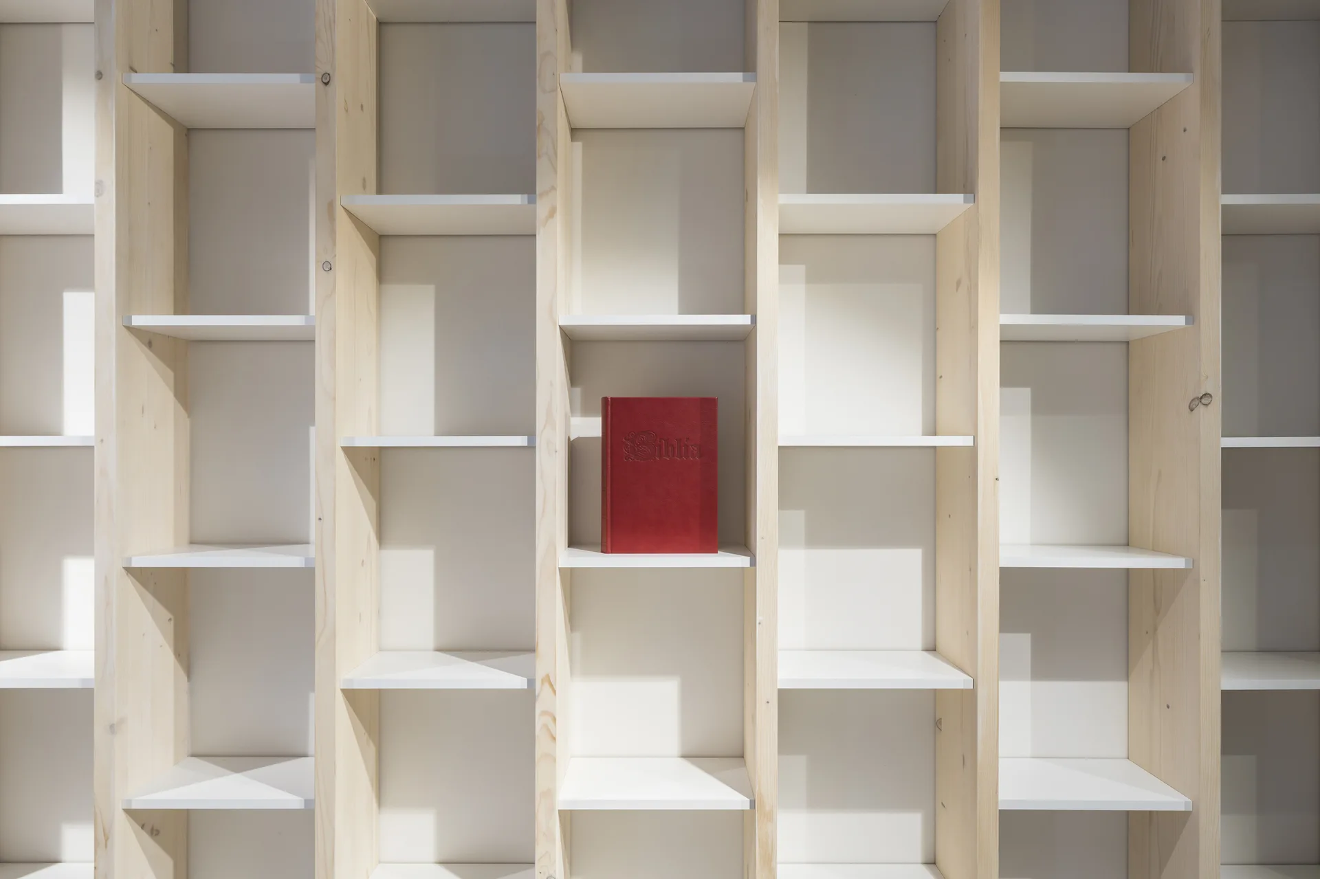 A custom light wood grid shelving unit features white horizontal shelves with a single red book placed in the center of one compartment.