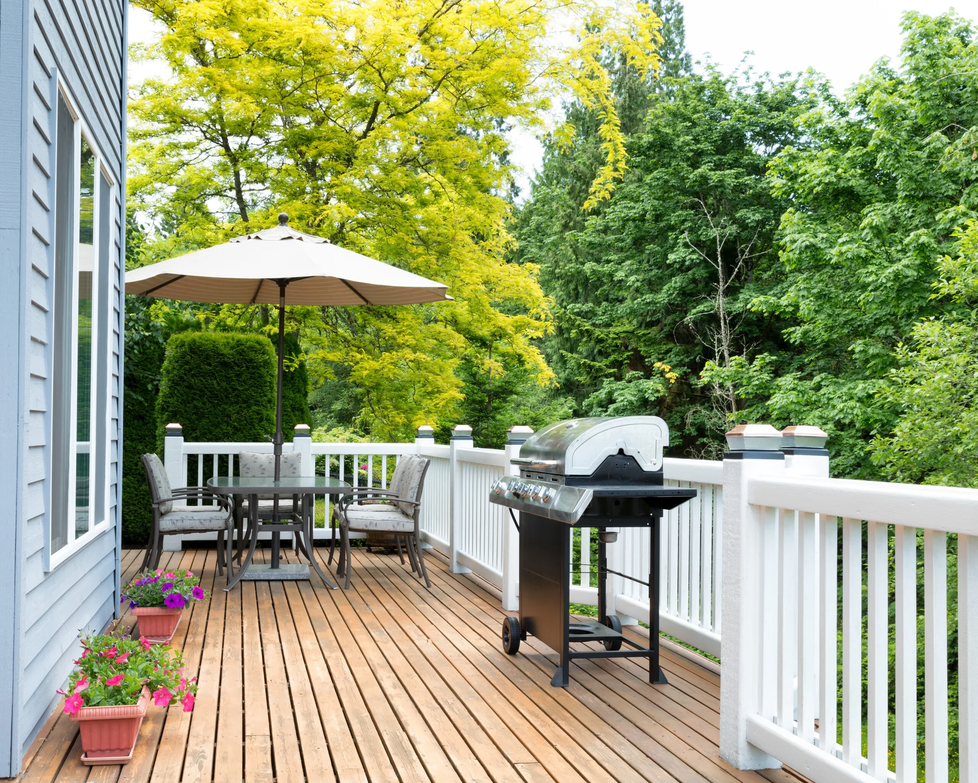 A wooden deck featuring a dining table set under a tan umbrella, a barbecue grill, white railings, and potted pink flowers against blue siding.