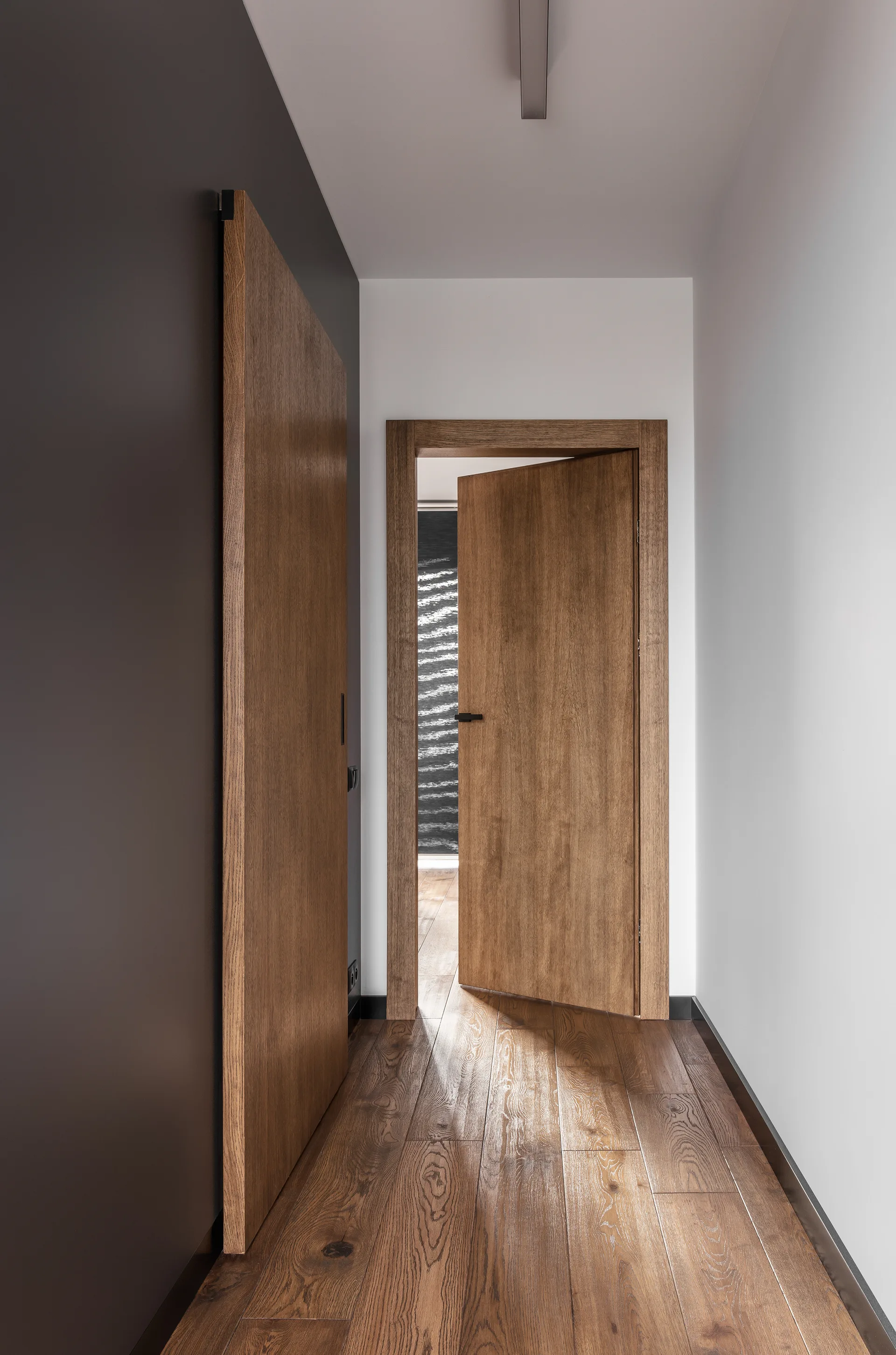 A modern residential hallway features warm wood-plank flooring, matching wooden doors, and contrasting dark grey and white walls.