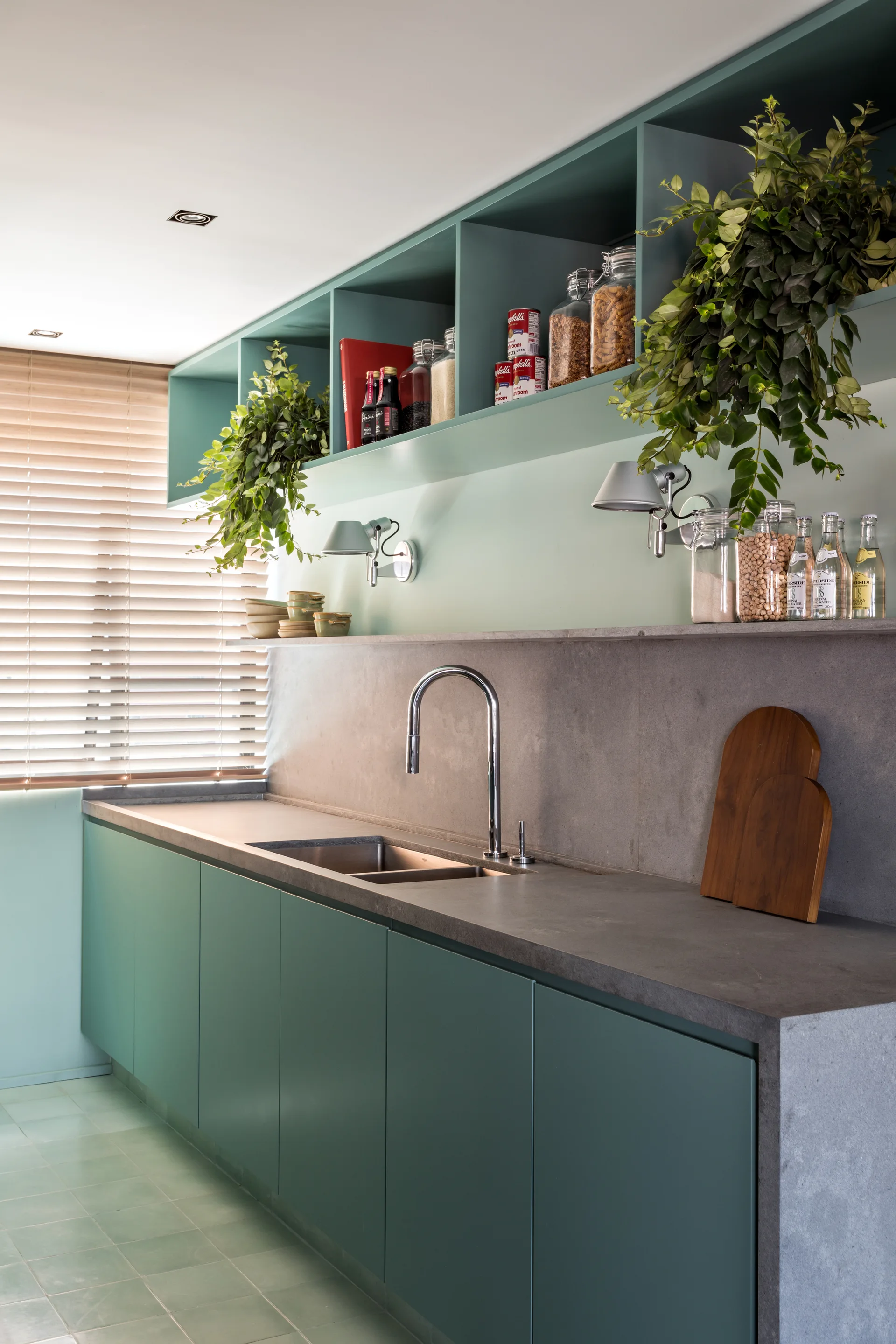 A modern kitchen features matte sage green cabinetry, a grey stone countertop and backsplash, a stainless steel sink, and open shelving.