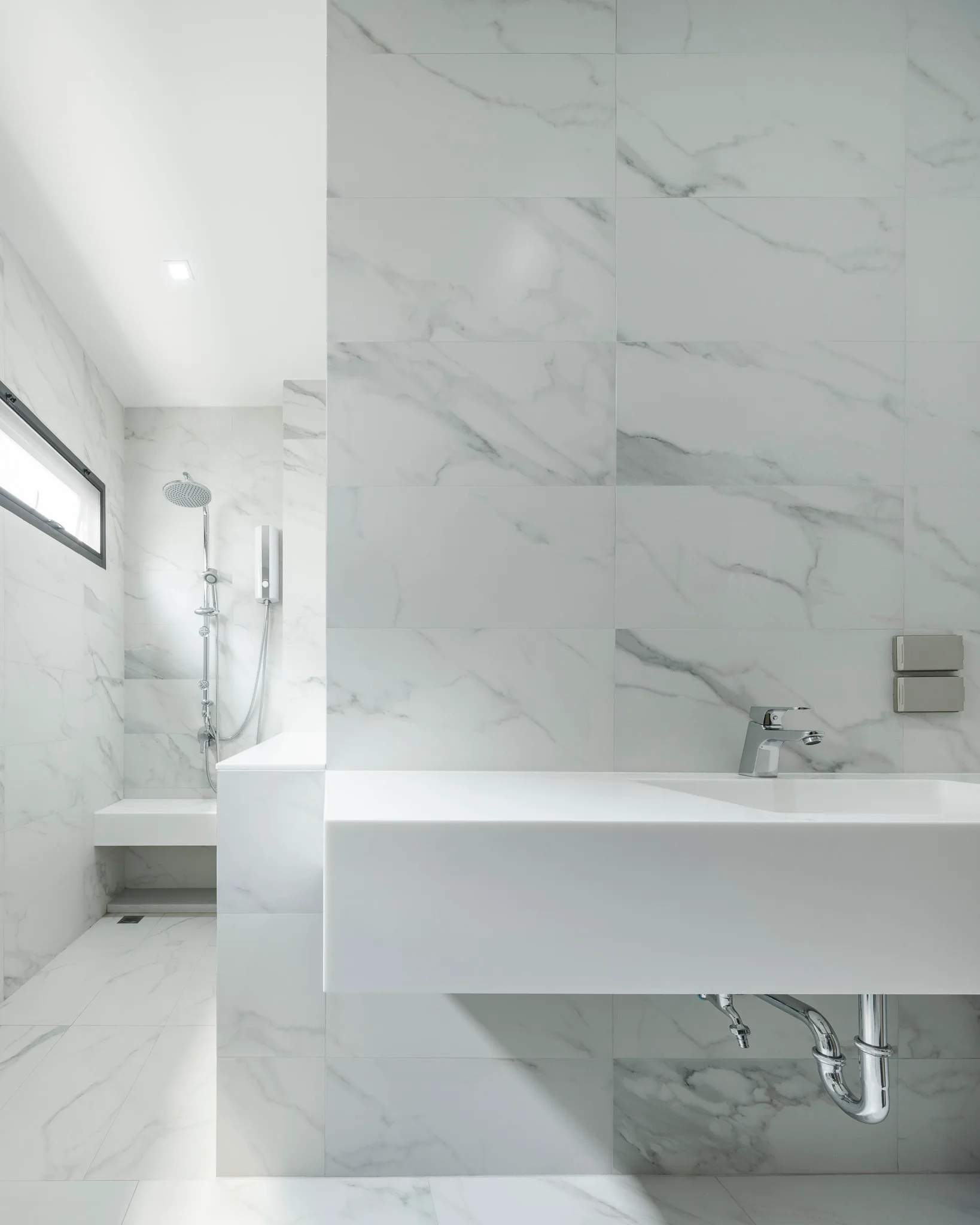 A modern white bathroom with large marble-patterned tiles, a wall-mounted sink, and a walk-in shower with a built-in bench.