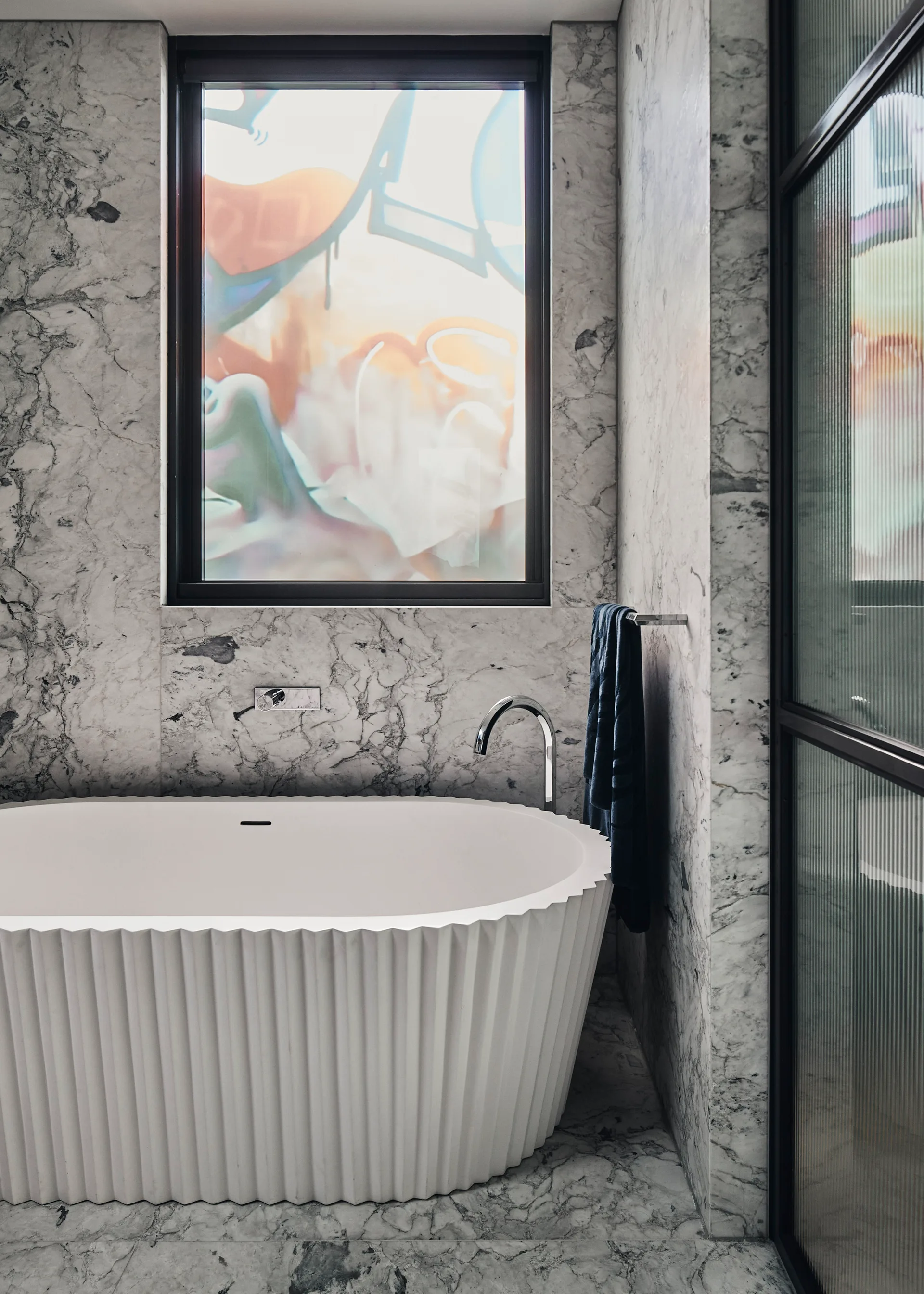 A white fluted freestanding bathtub sits in a bathroom with heavily veined gray and white marble walls and a window.