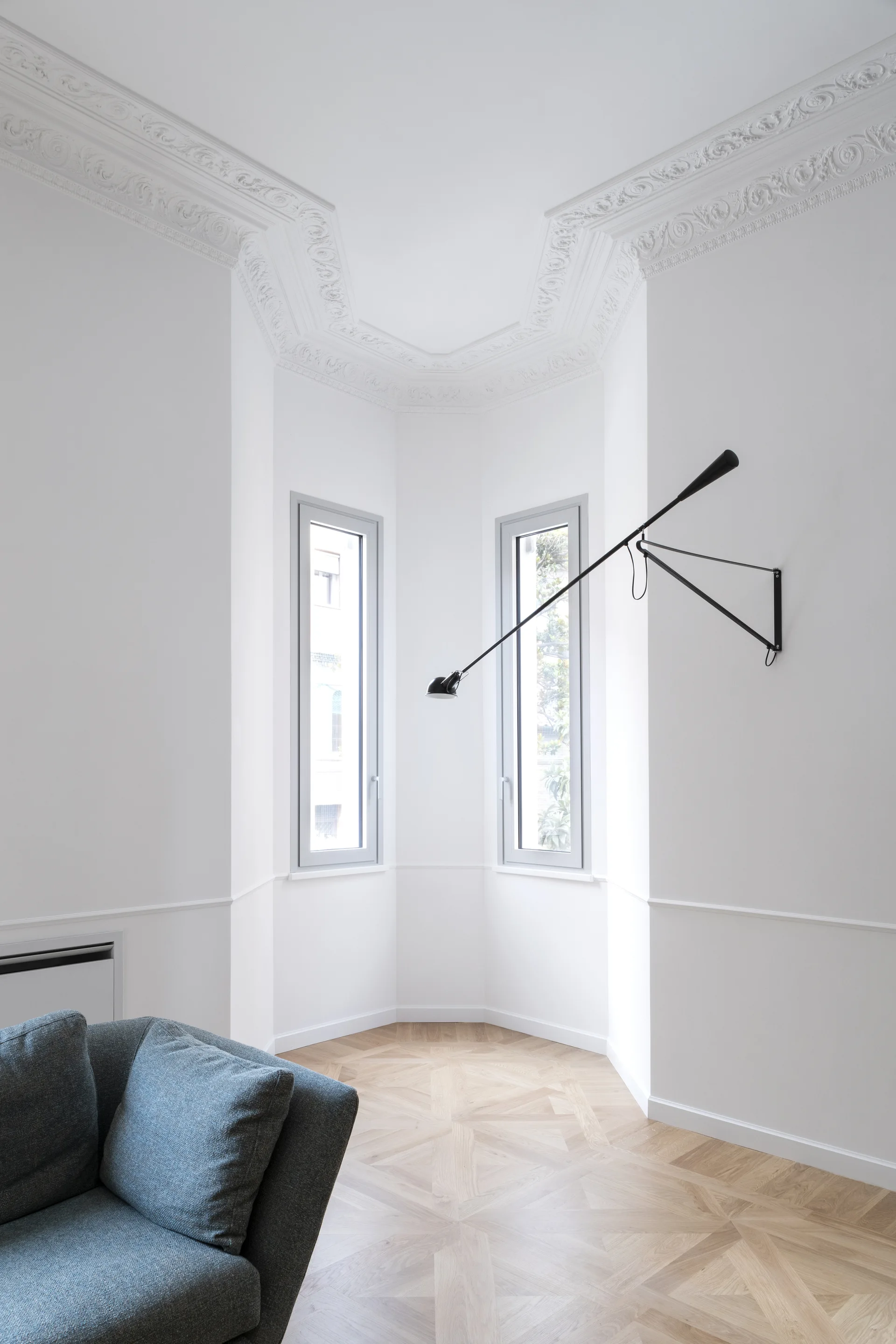 A bright white living room corner features parquet wood flooring, ornate ceiling crown molding, a grey sofa corner, and a black articulating wall lamp.