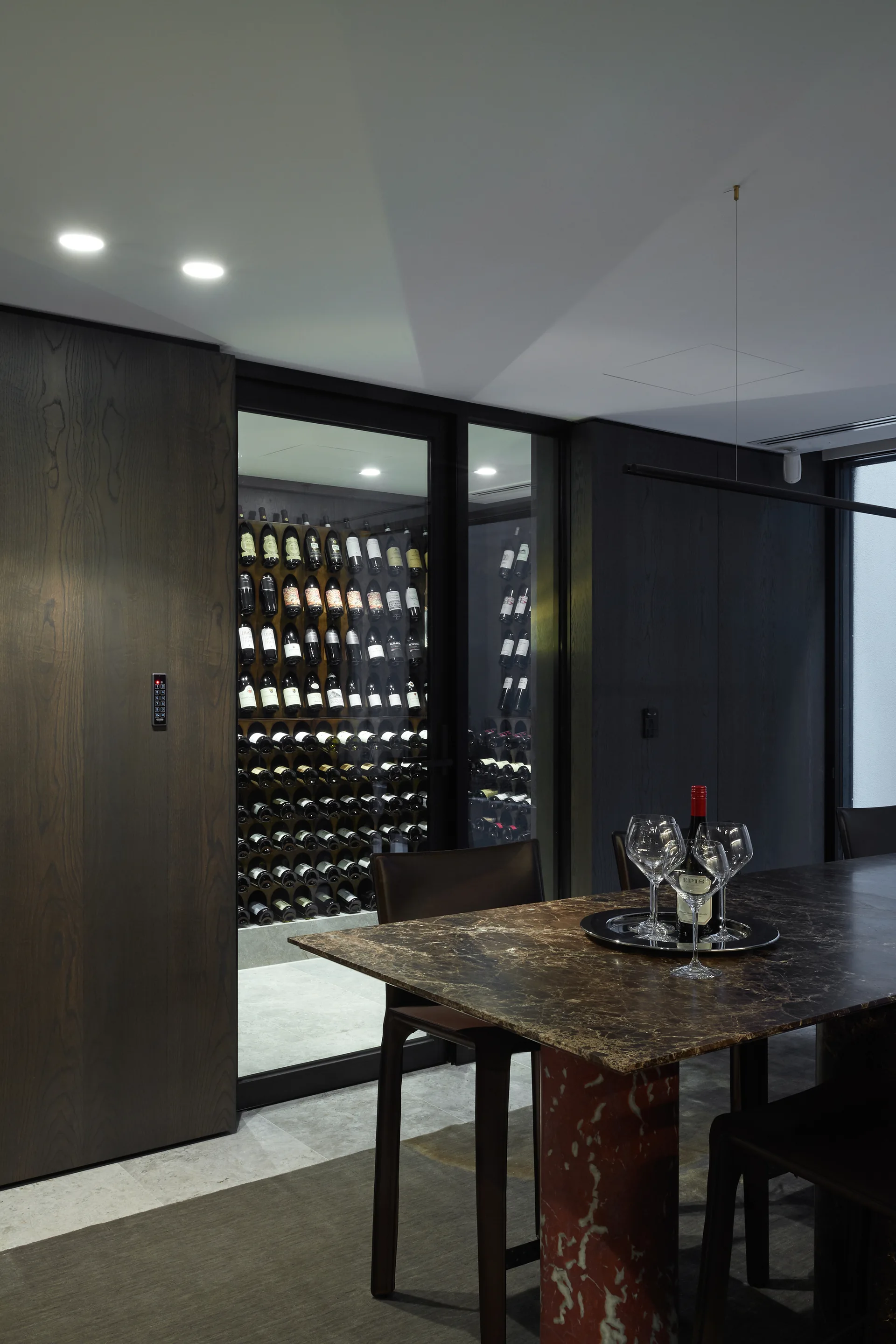 A moody dining room features a veined stone dining table next to a glass-enclosed wine room with illuminated storage racks.