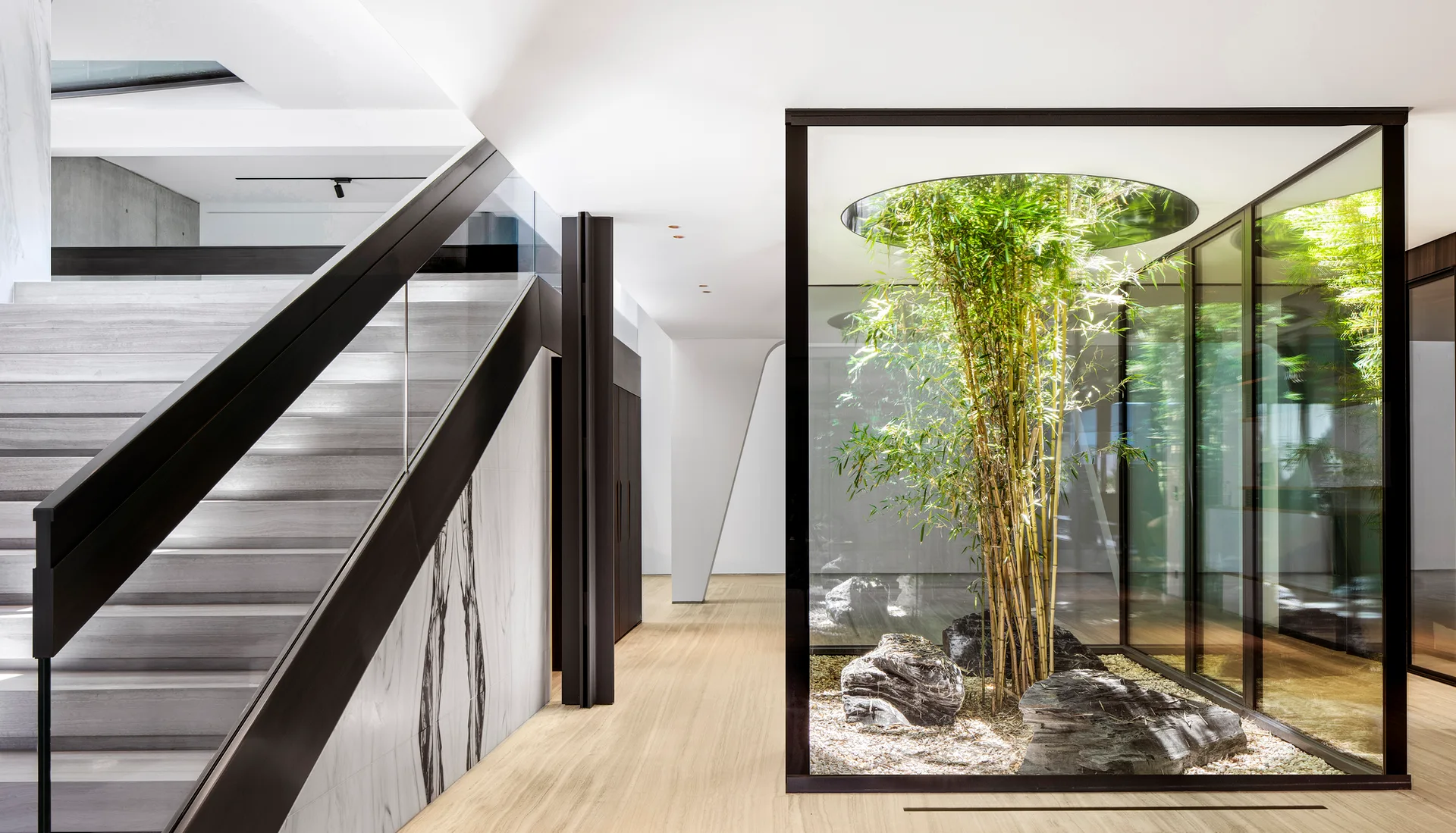 A minimalist hallway featuring a wide stone staircase beside a glass-enclosed indoor courtyard with green bamboo and natural stones.
