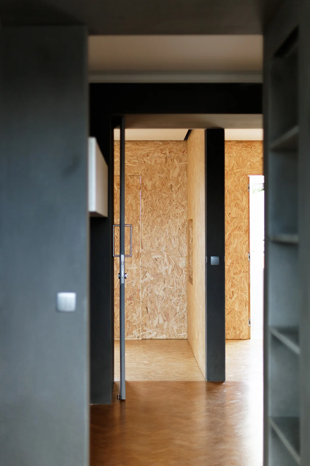 A view through a dark grey doorway into an interior hallway featuring walls and flooring clad in textured oriented strand board paneling.