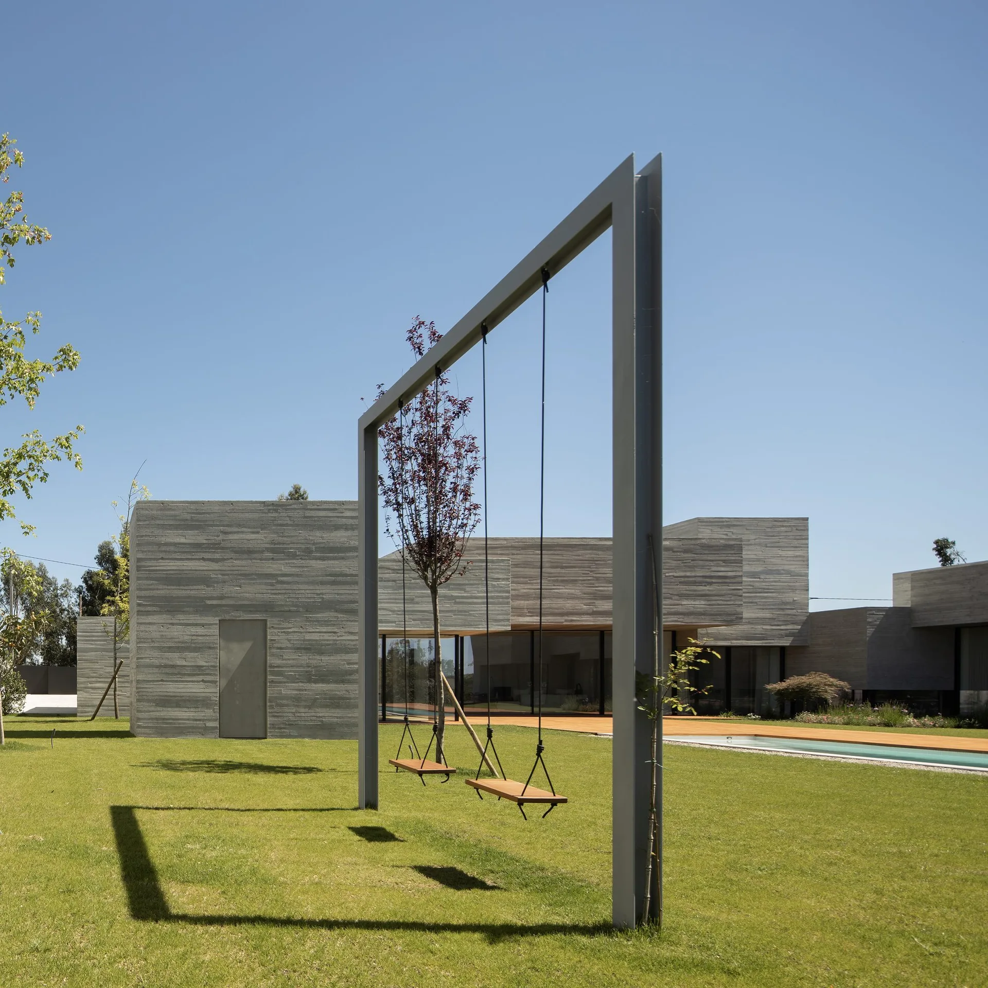 A tall gray metal swing frame stands on a green lawn in front of a modern concrete house with a swimming pool.