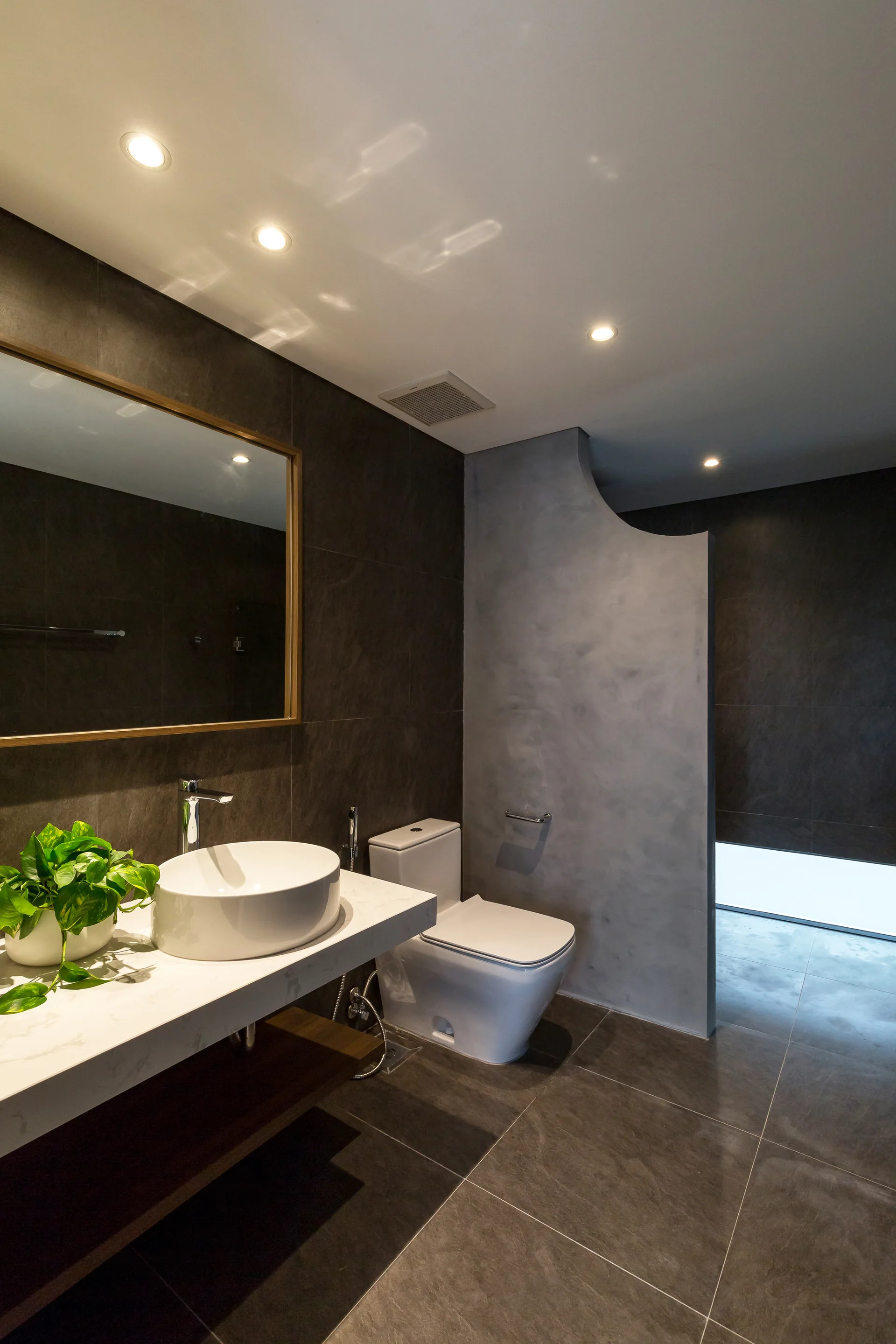 A modern bathroom features a white vessel sink on a floating vanity, a toilet, a curved grey partition, and dark grey tiled walls.