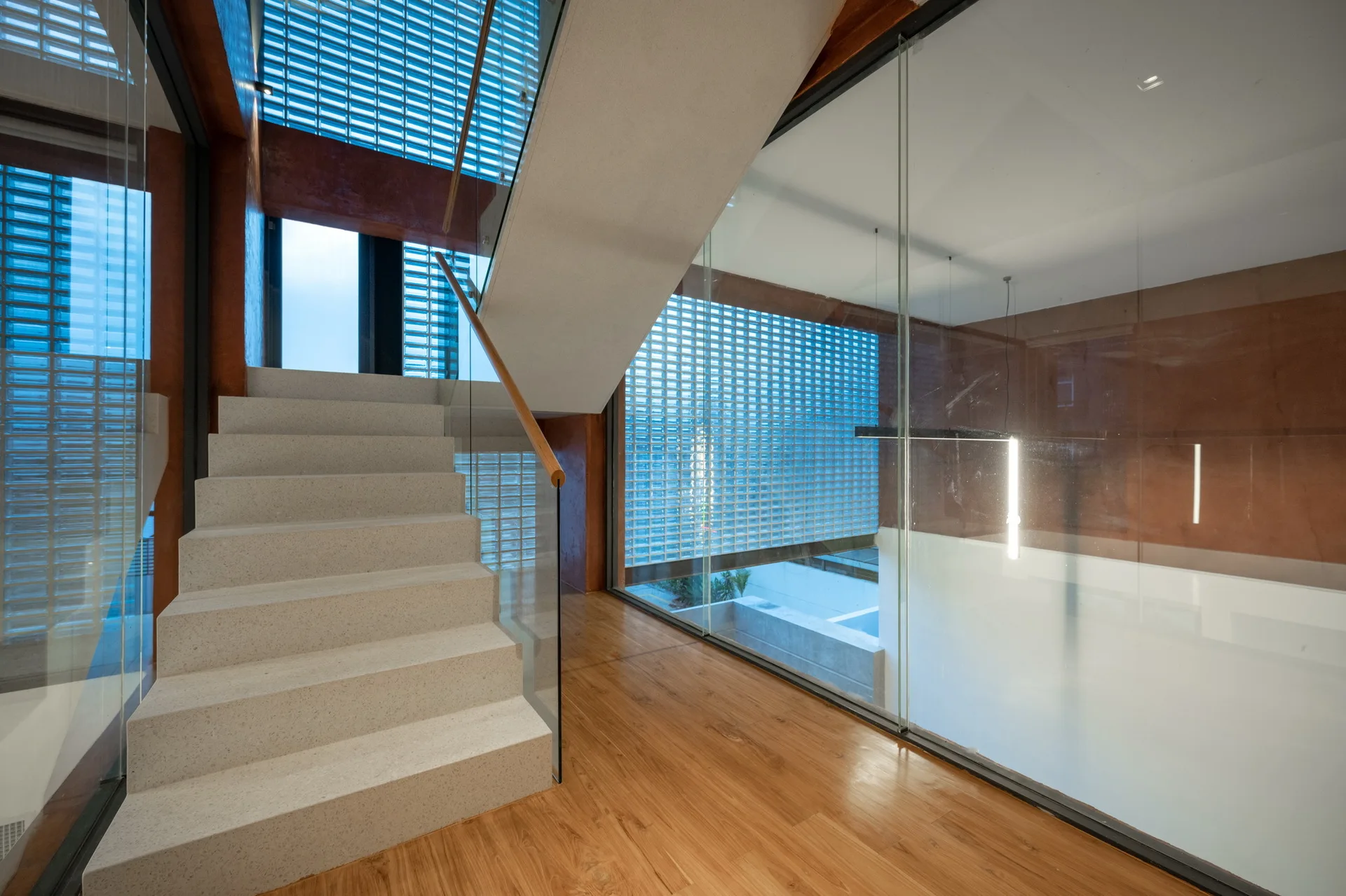 A light-colored concrete staircase with a glass railing stands next to a wooden floor and a large translucent glass block wall under a white ceiling.