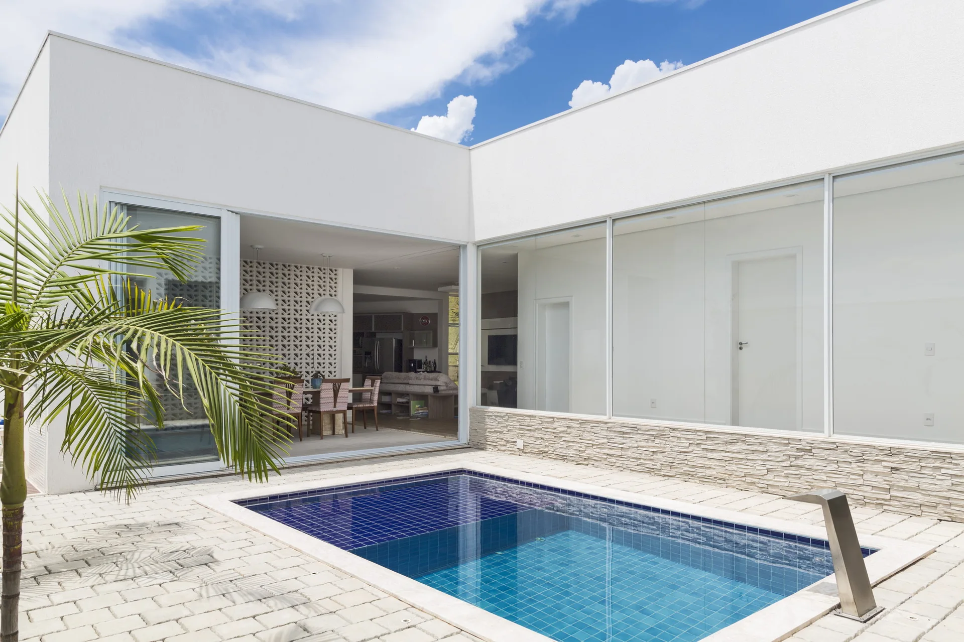 A rectangular swimming pool with blue tiles on a stone paved patio next to a white house with large sliding glass doors showing an indoor dining area.