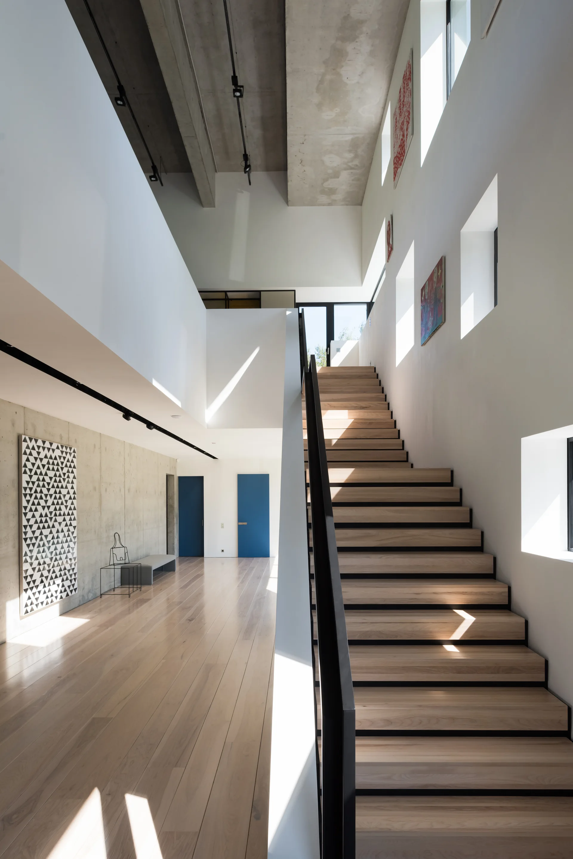 A modern wood-tread staircase with black metal railing rises next to a white wall with square windows, an exposed concrete ceiling, and blue accent doors.