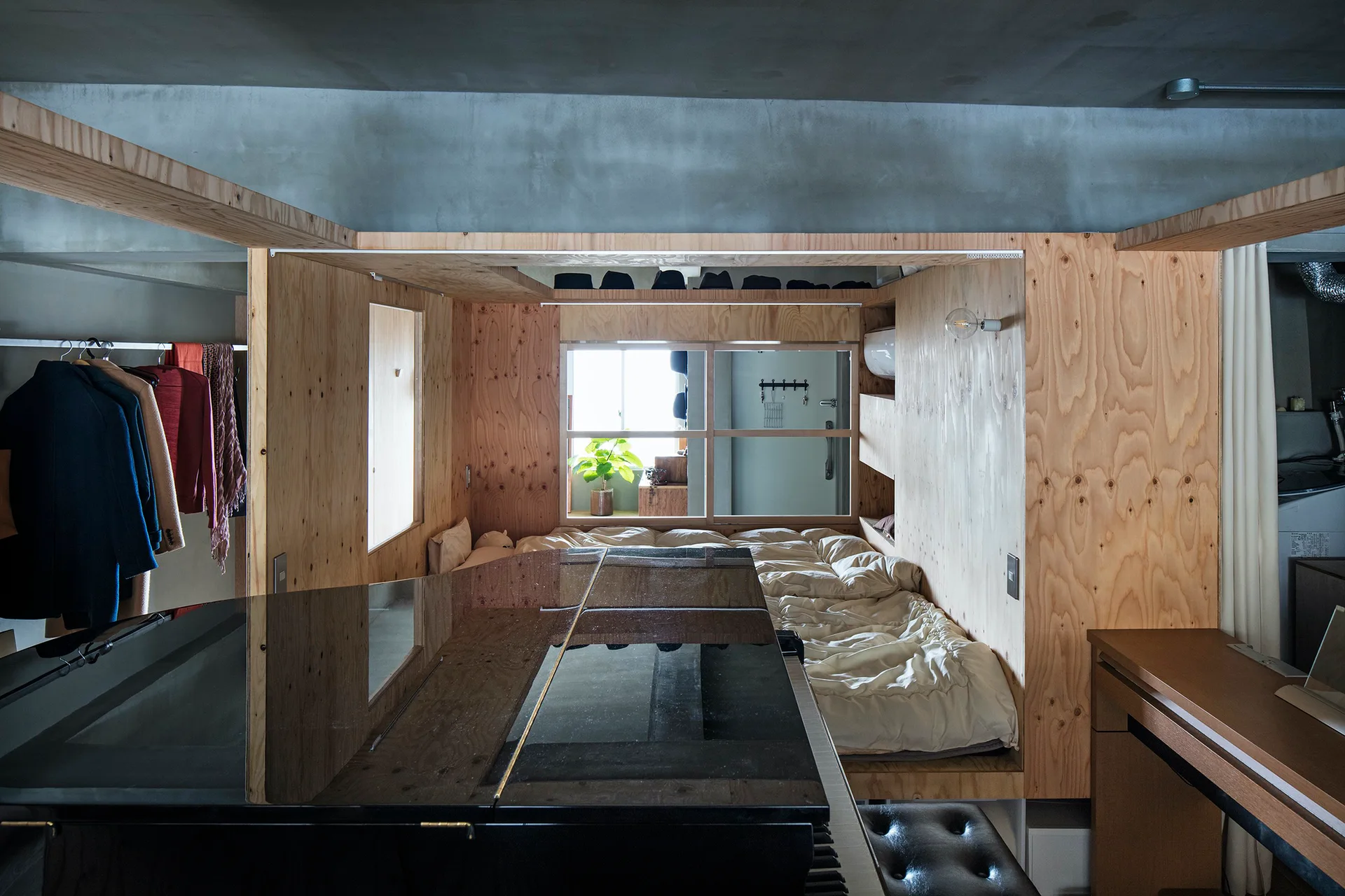 A loft apartment features a custom plywood sleeping box enclosing a bed next to a black grand piano under an exposed concrete ceiling.