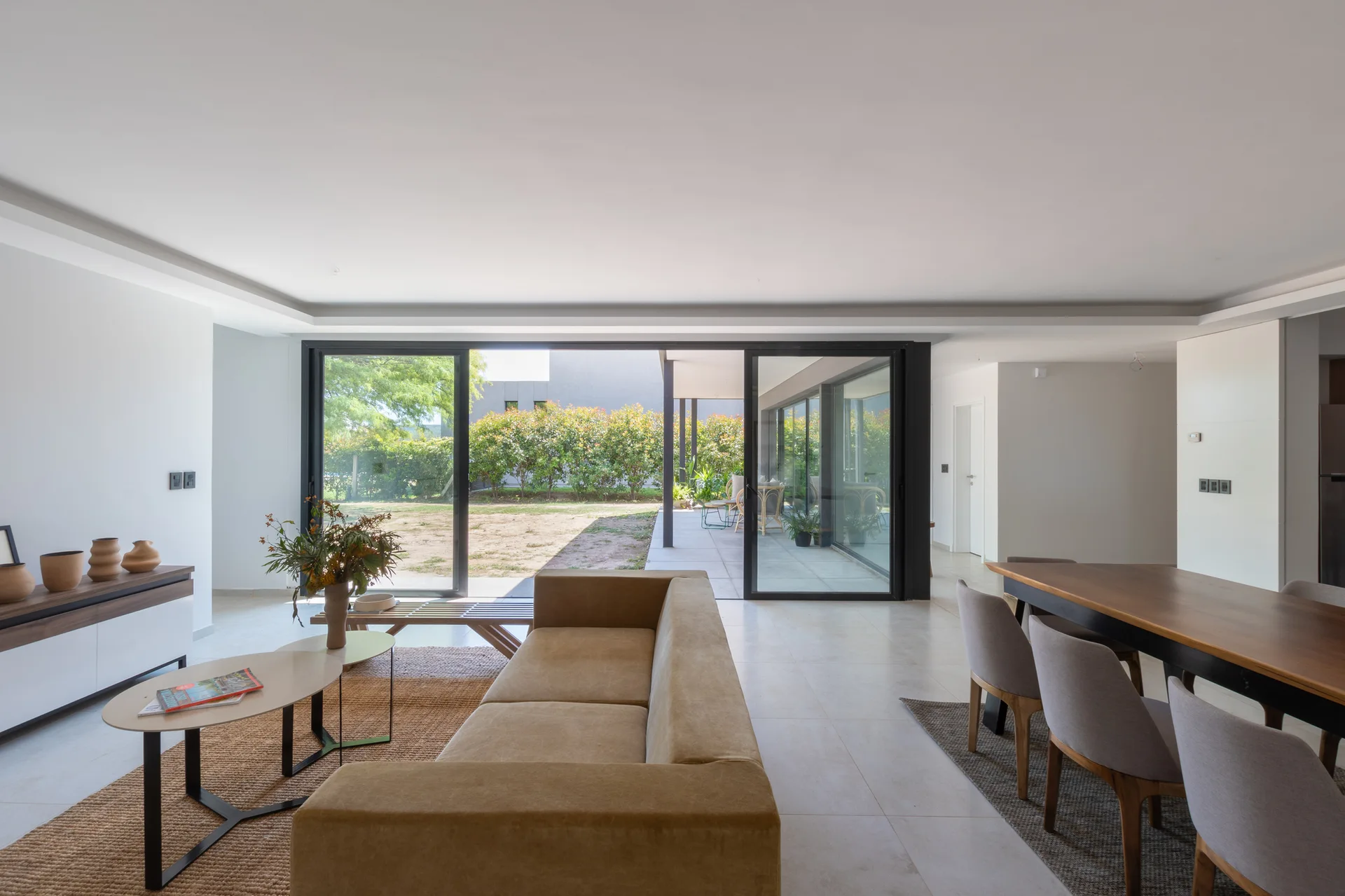 An open-concept living and dining room with a beige sofa, a wood dining table, and large sliding glass doors leading to a garden.