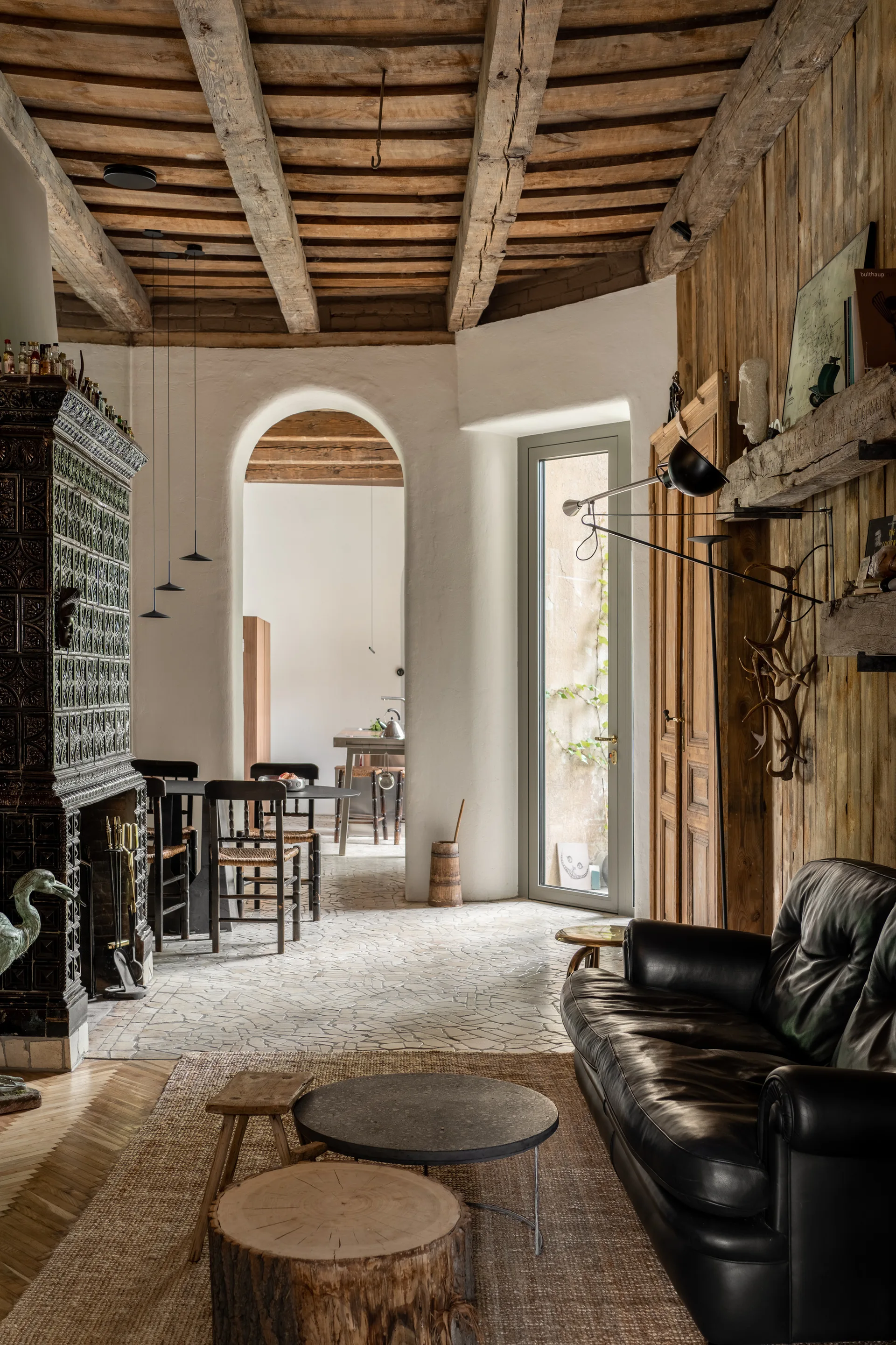 A rustic living room features an ornate green tiled fireplace, a black leather sofa, exposed ceiling beams, and a stone mosaic floor.