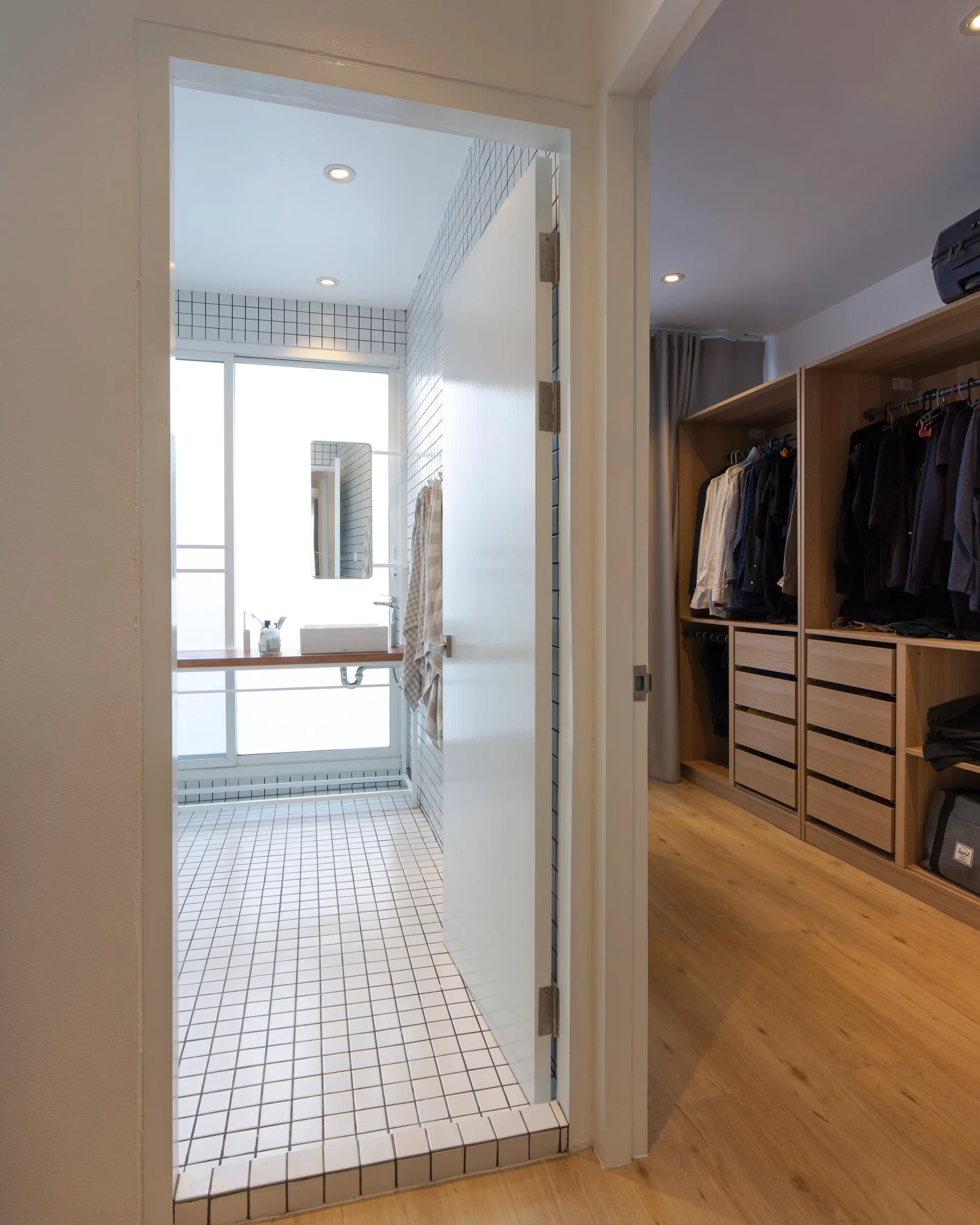A bathroom featuring white grid-tiled walls and floors with a floating wood vanity next to an open walk-in closet with wooden cabinetry.