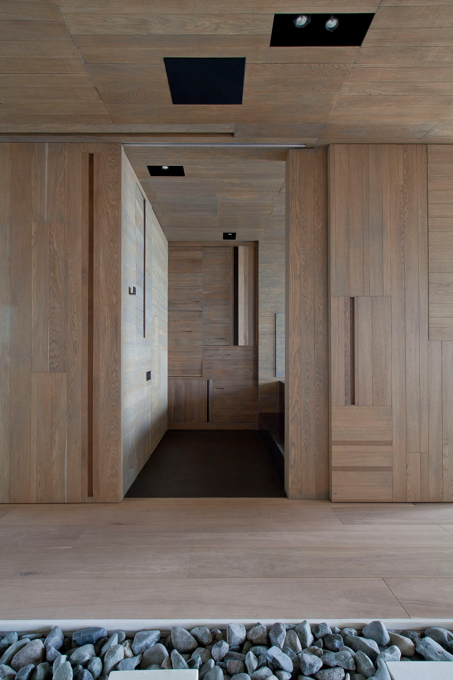 A modern hallway with light wood paneling on the walls and ceiling, flush cabinetry, black recessed lights, and grey river stones in the foreground.