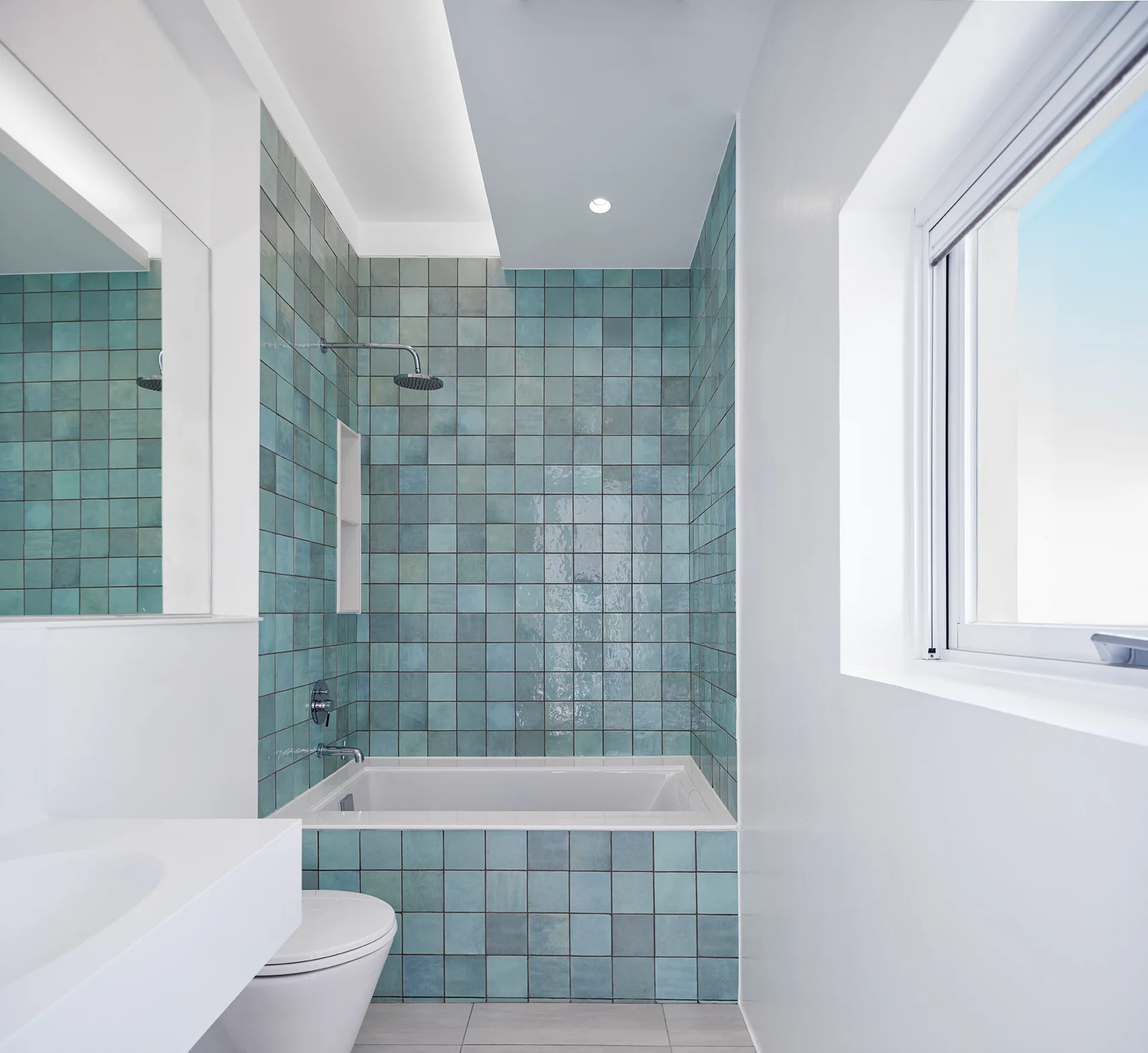 A minimalist bathroom features a built-in bathtub surrounded by glossy teal square tiles, a white floating vanity, and a large window.