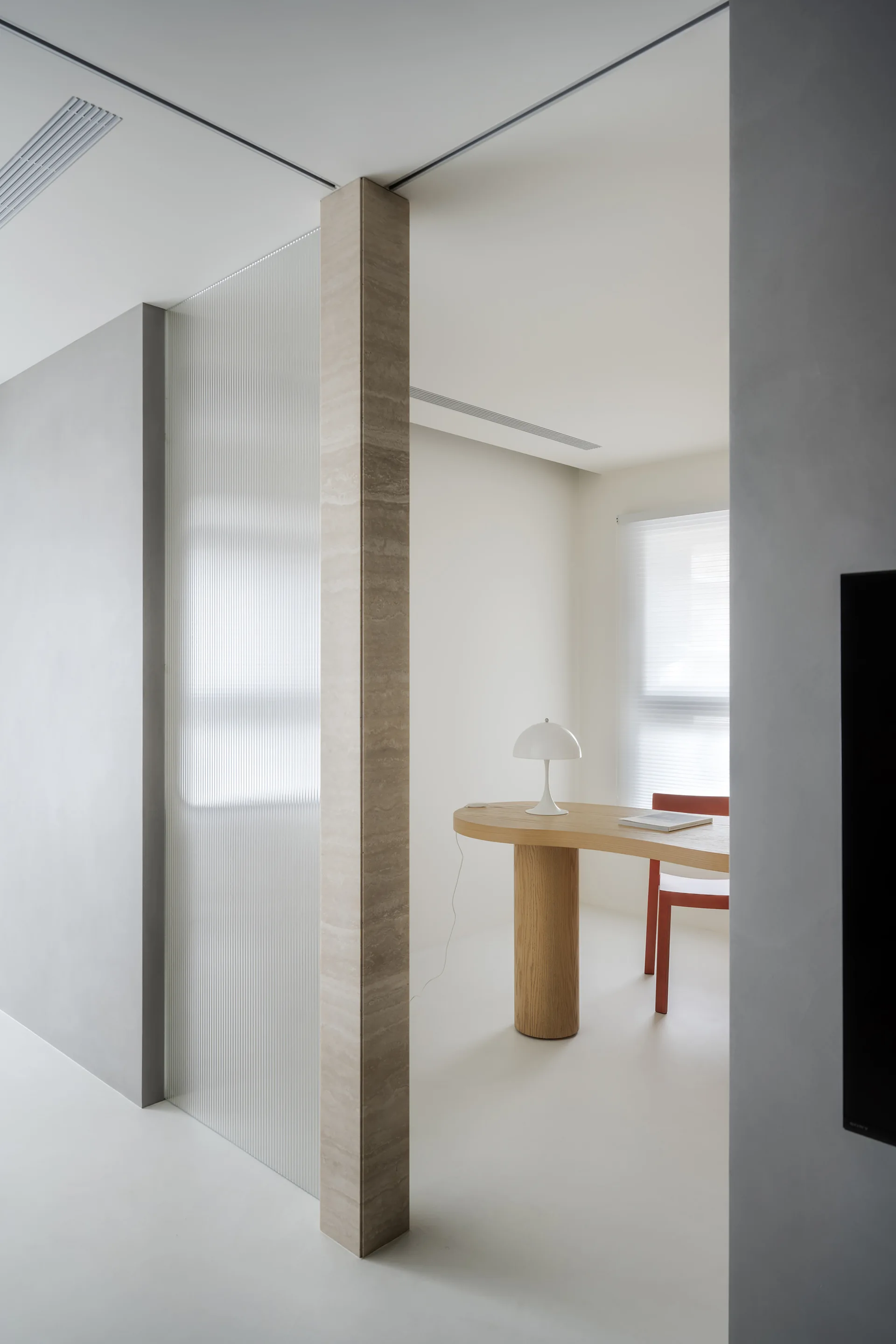 A minimalist home office features a curved wooden desk, a red chair, and a translucent fluted glass partition supported by a stone pillar.