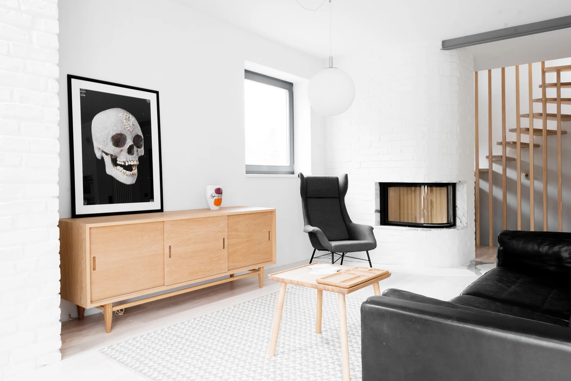 A modern living room features a light wood sideboard with a framed skull print, a black leather sofa, and a corner fireplace against a white brick wall.
