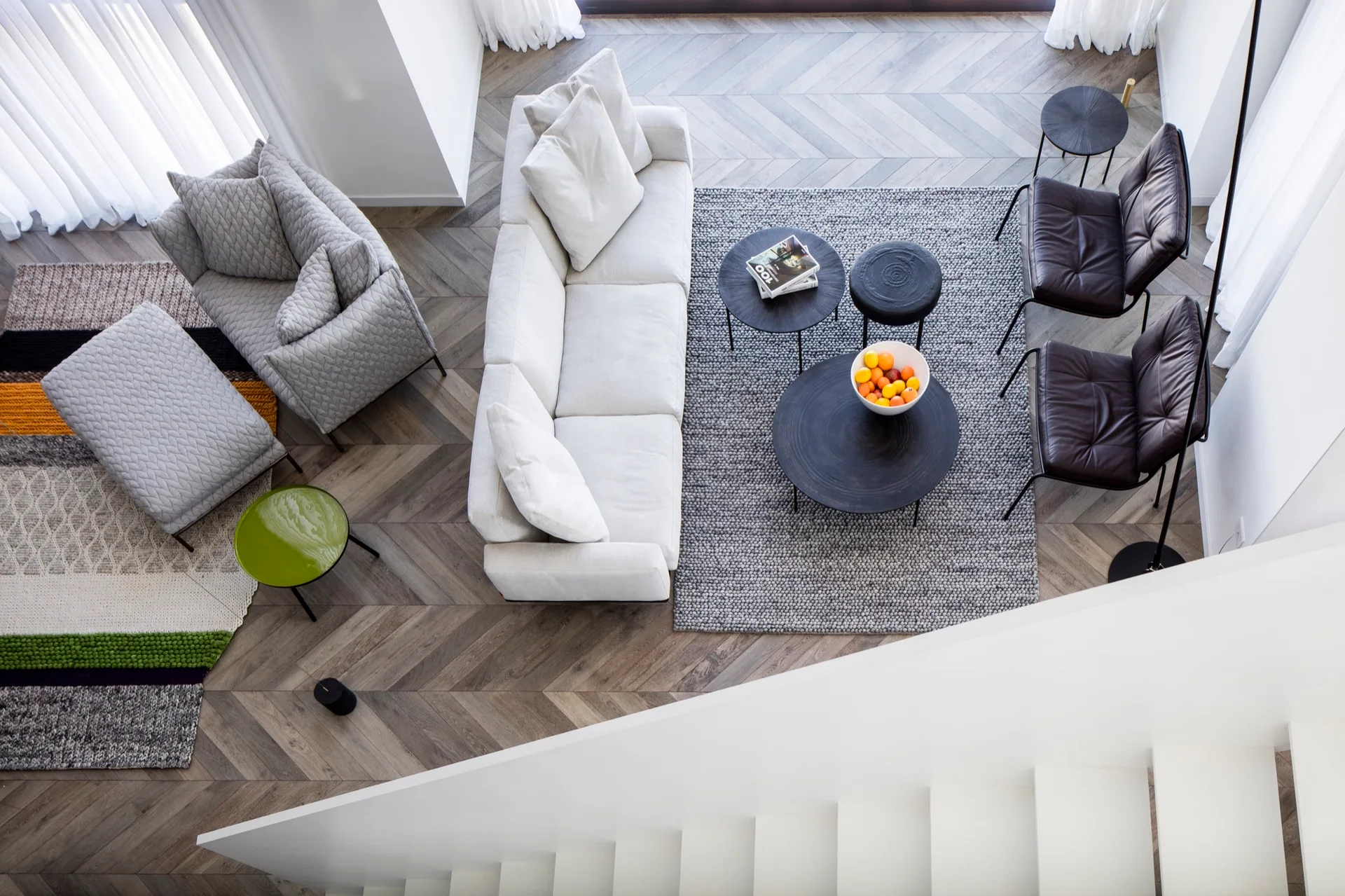 An overhead view of a modern living room featuring a long white sofa, gray quilted armchairs, black coffee tables, and herringbone wood floors.
